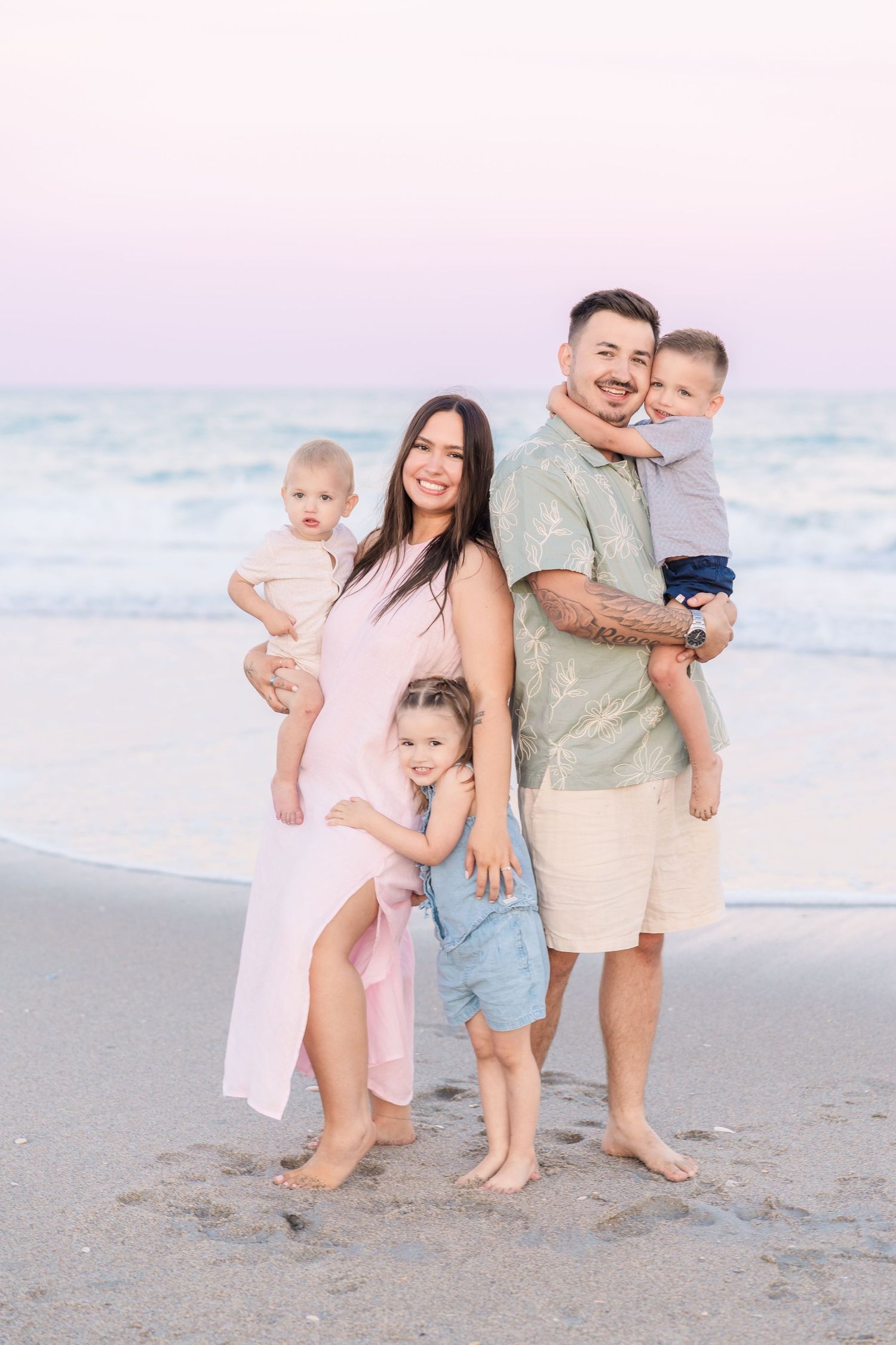 Family of five poses on beach at sunset, parents holding babies while toddler stands between them.