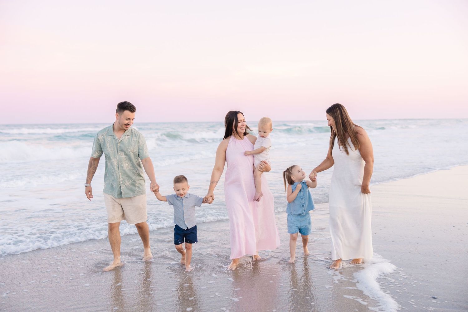 Family of five walking along a sandy beach at sunset, with parents and children holding hands by the ocean waves.