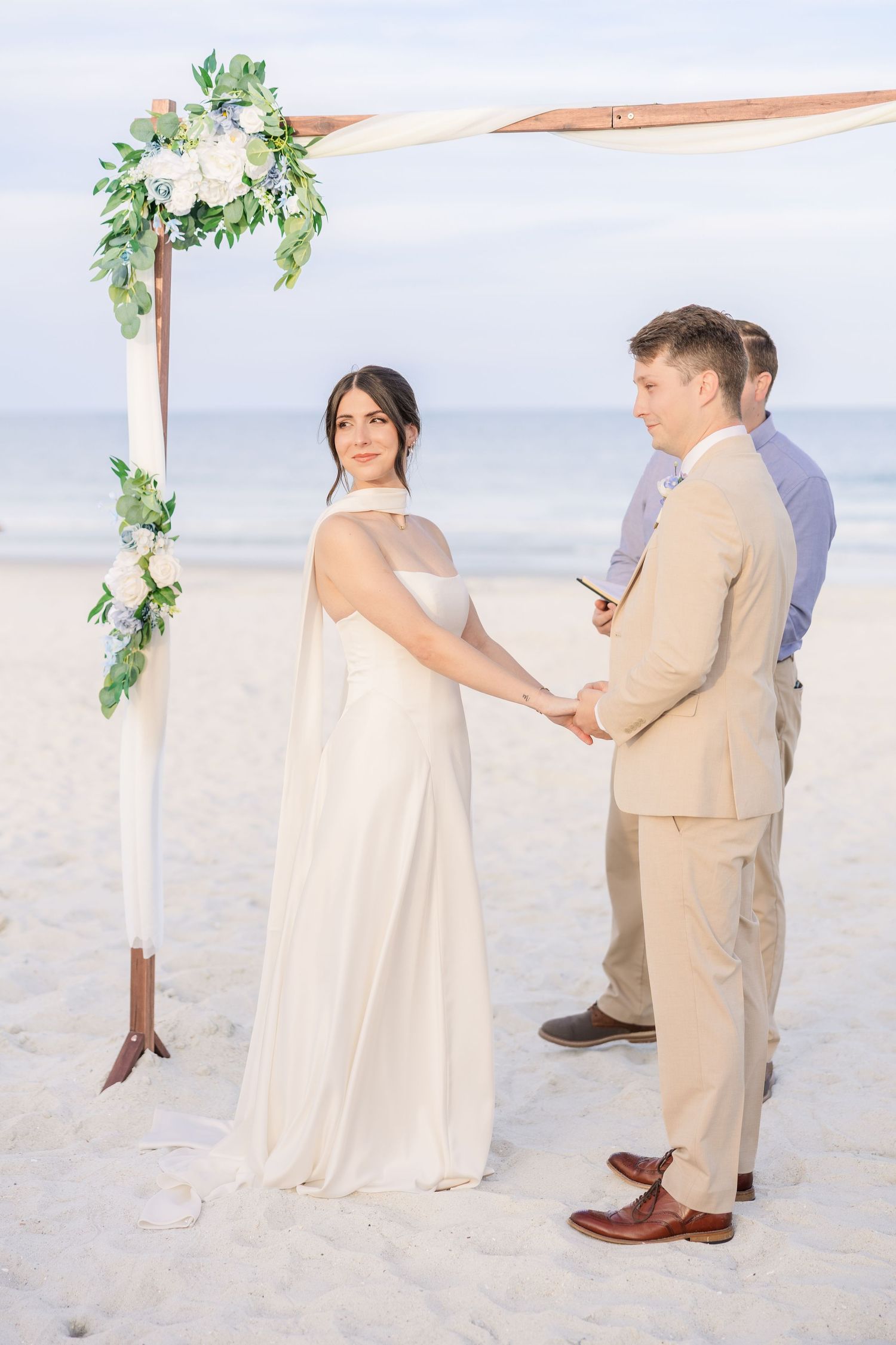Bride and groom holding hands at beach wedding altar decorated with white flowers and greenery in Carolina Beach near Wilmington, NC.