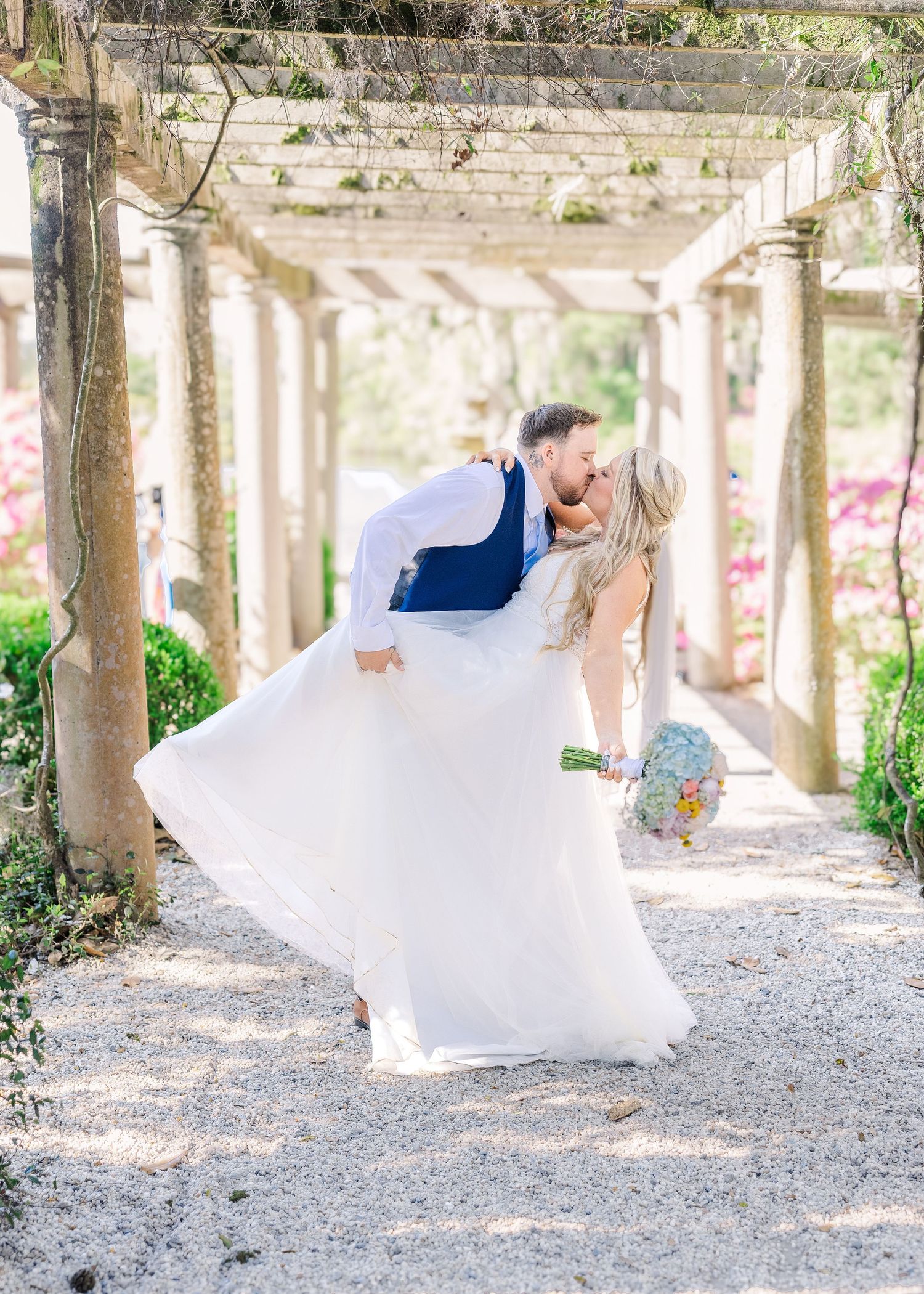 Groom dips bride for a kiss under a garden pergola, her white gown flowing as she holds a colorful bouquet in Airlie Gardens in Wilmington,