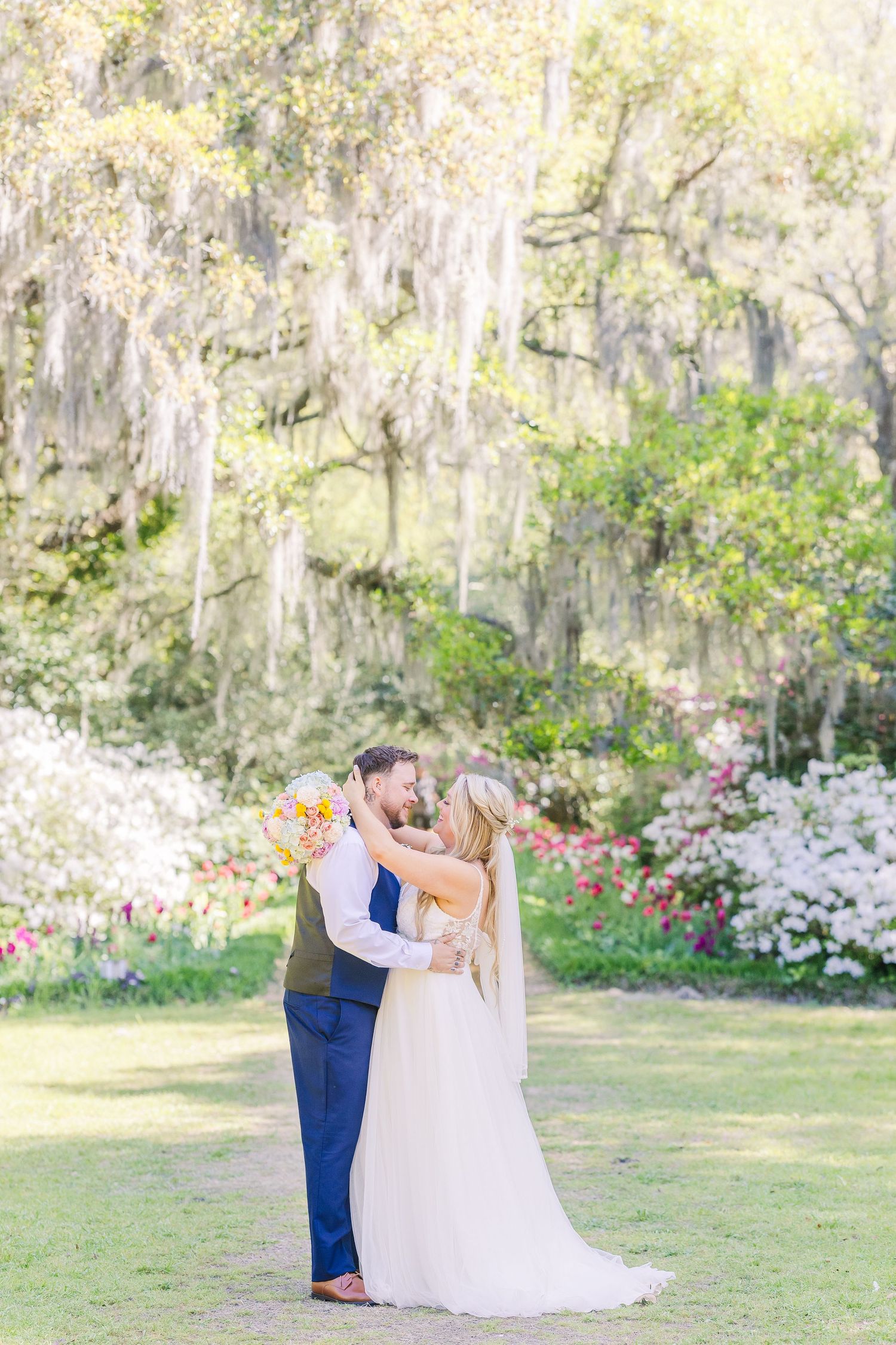 Bride and groom share a romantic moment in a lush garden with Spanish moss-draped trees and colorful blooms in Airlie Gardens in Wilmington,