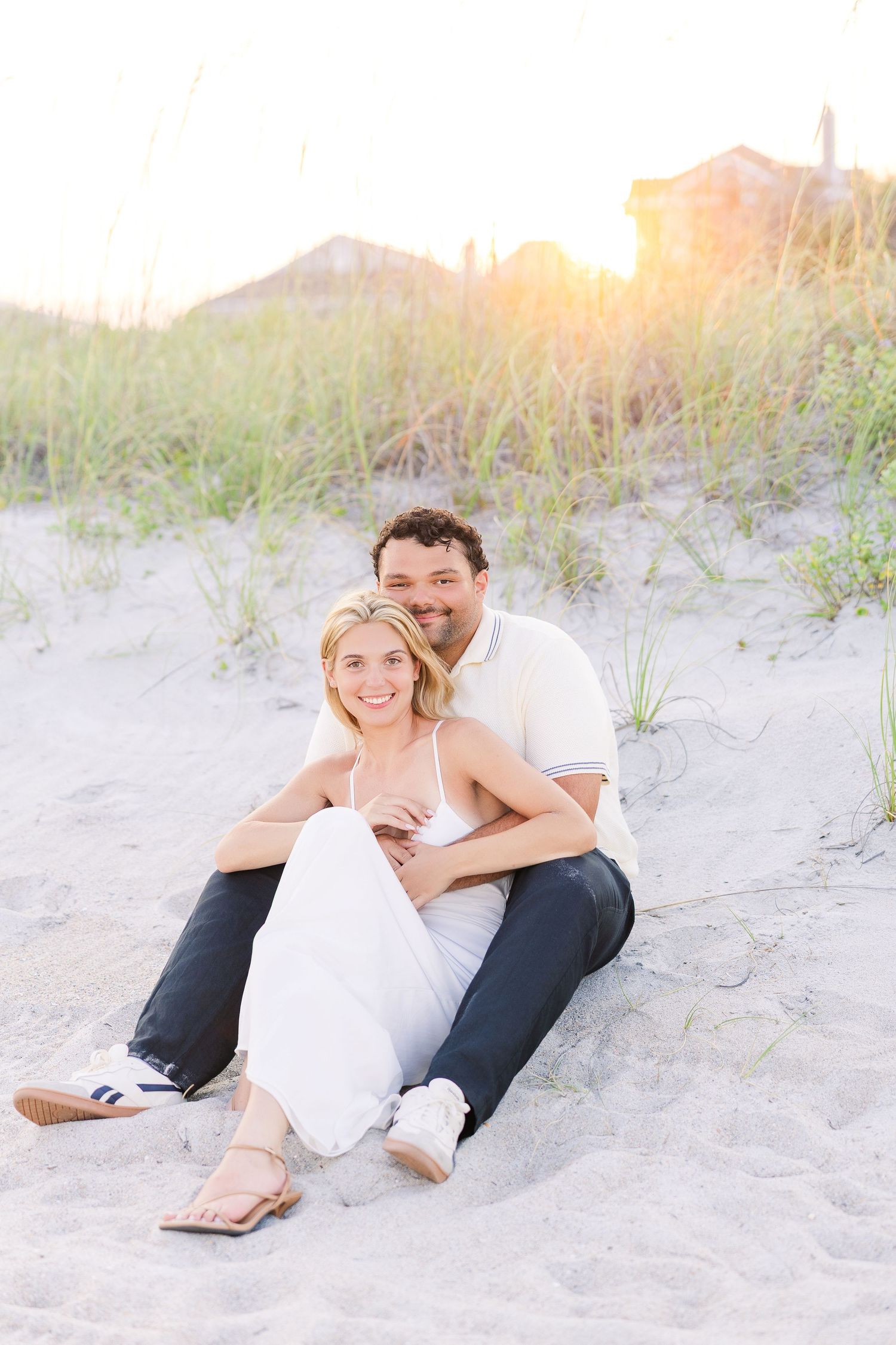 Smiling couple sits together on sandy beach dunes at sunset in casual white outfits.