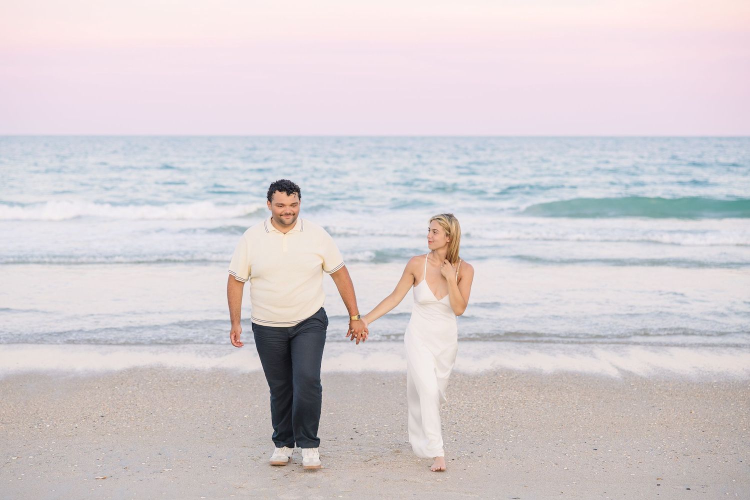 Couple walking hand in hand along a beach at sunset with pastel pink and blue sky over the ocean.