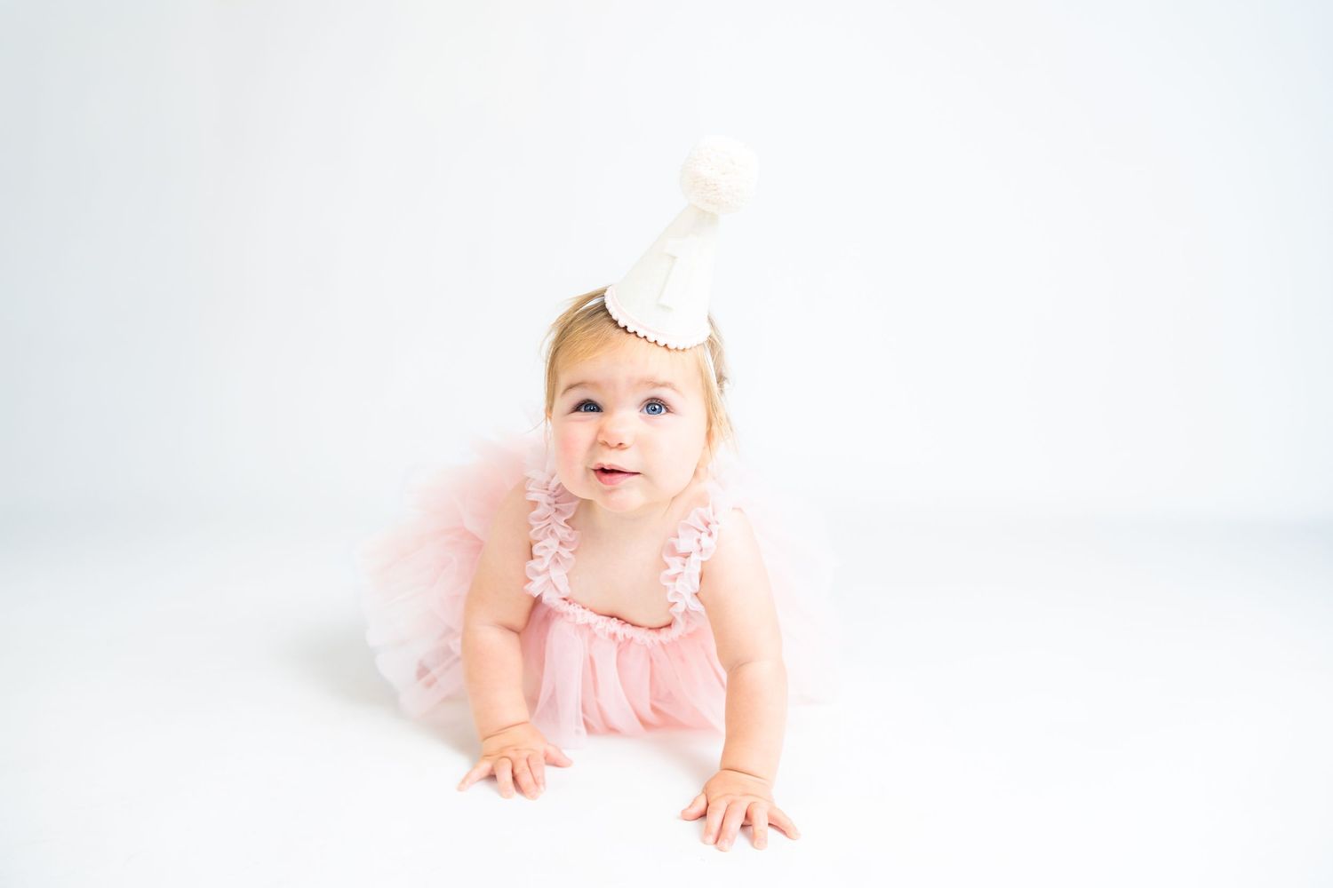 Baby in pink tulle dress crawls on white floor during studio photo session.