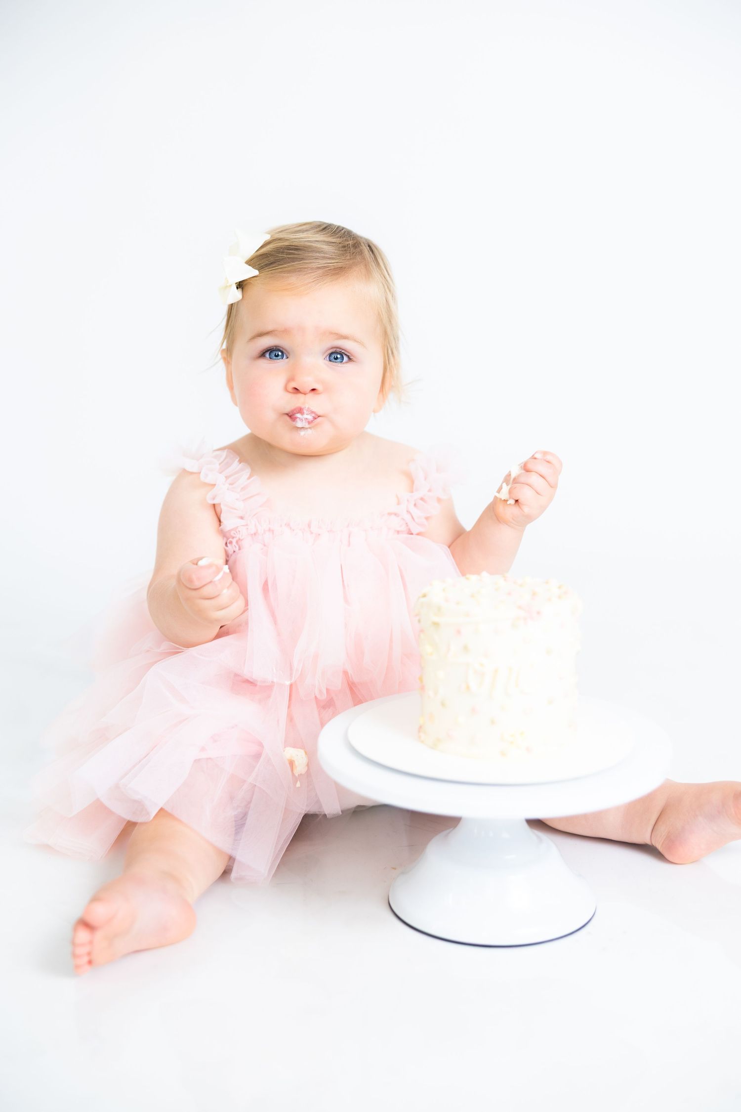 Baby in pink tulle dress sits next to white cake stand against white background.