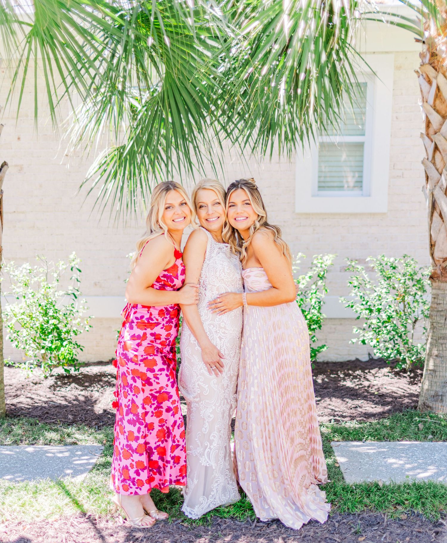 Three women in elegant dresses pose together under palm trees against a light colored building.
