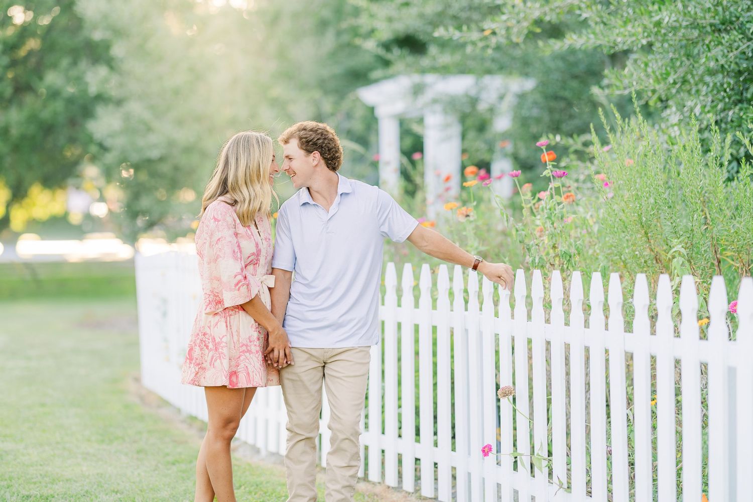 A romantic couple strolls along a white picket fence on a sunny day in a garden setting.