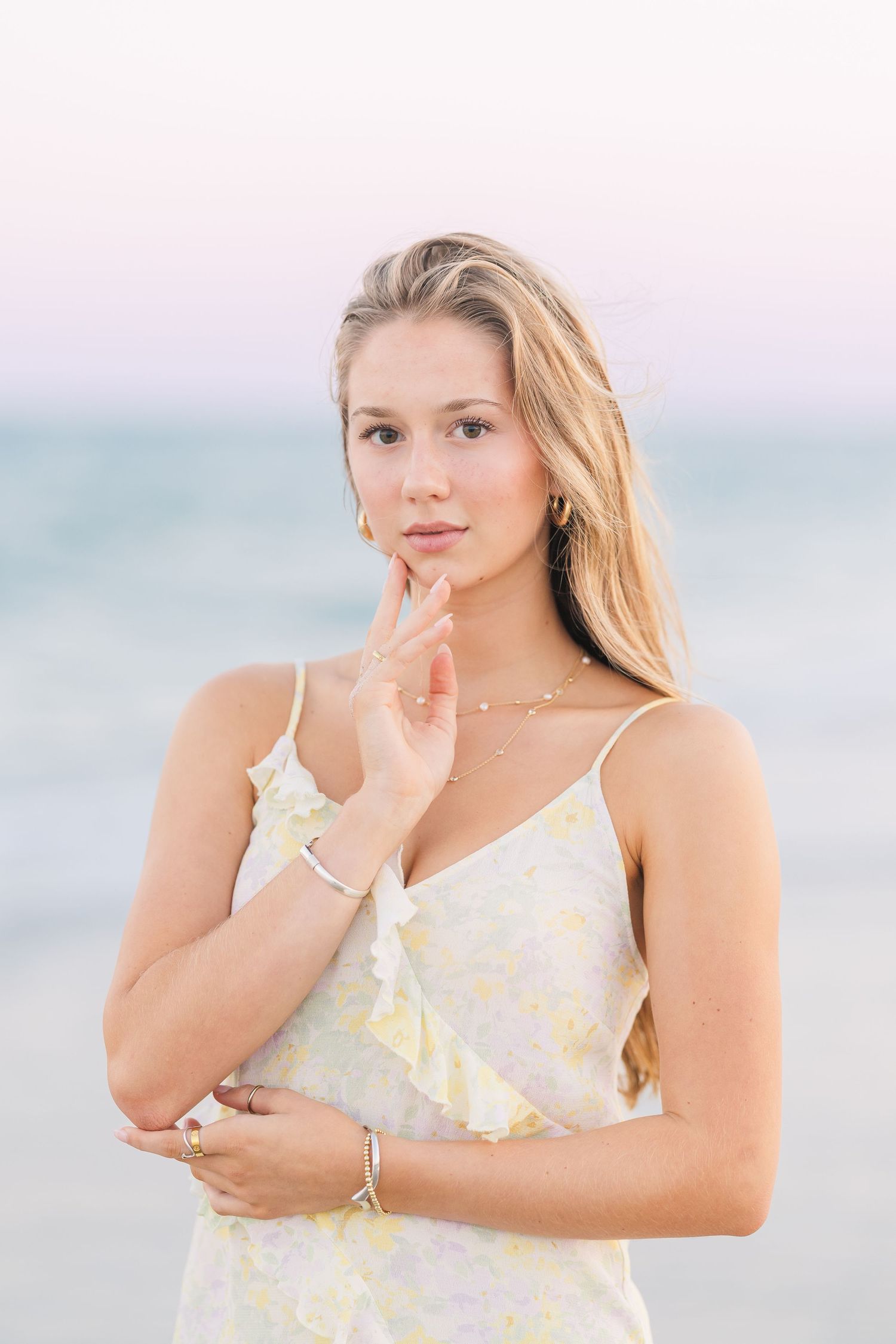 Woman in yellow floral dress poses gracefully against pastel beach backdrop.