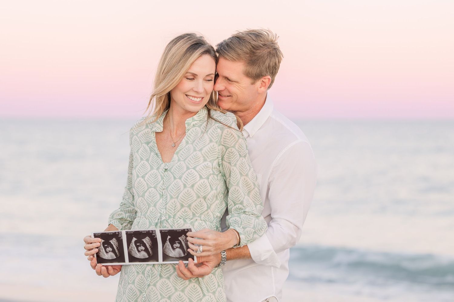 Couple holding ultrasound photos during sunset beach pregnancy announcement.