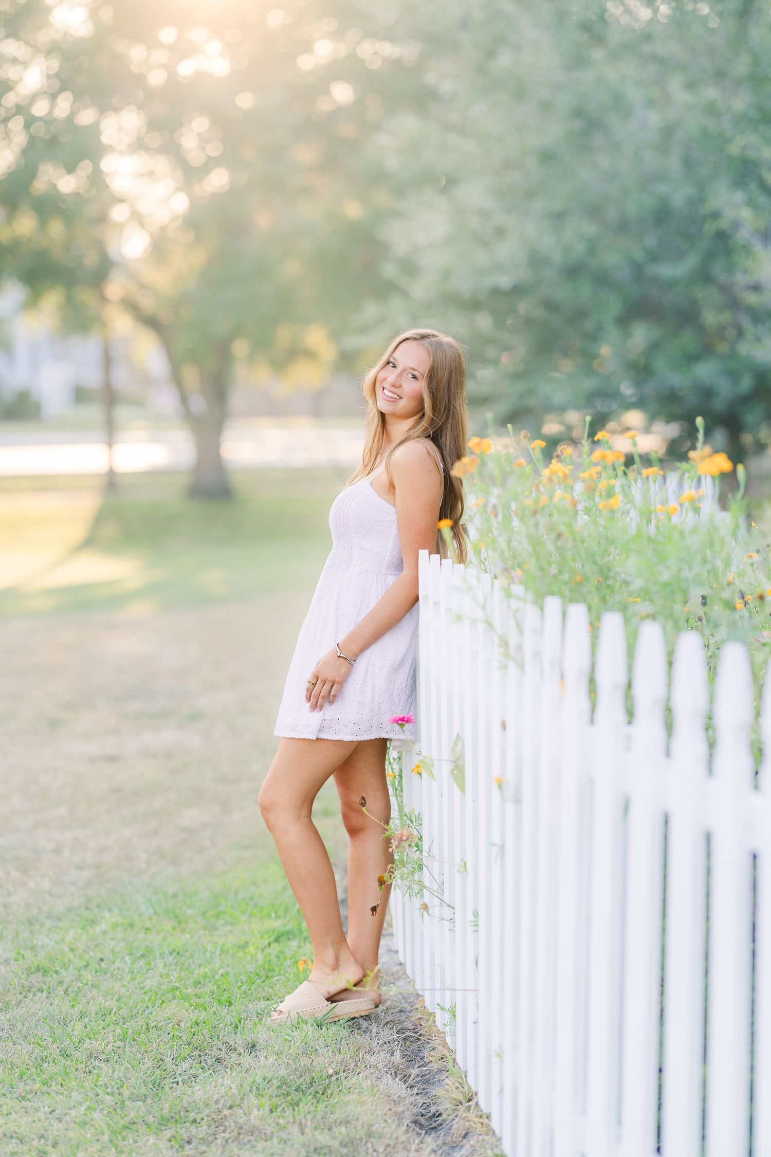 Figure in white dress plays near white picket fence surrounded by summer flowers.
