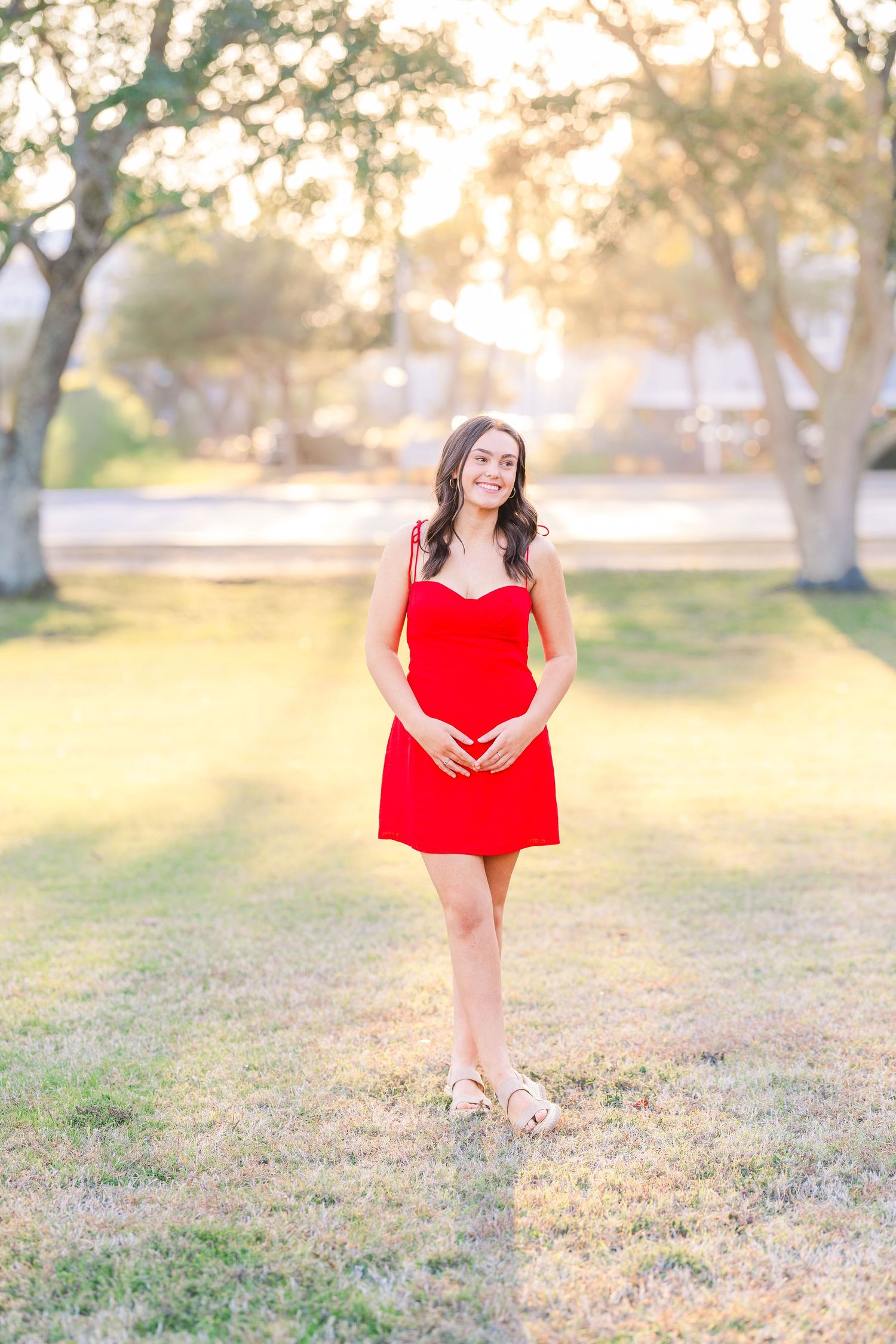 Golden hour portrait in red sundress standing on grass with trees.