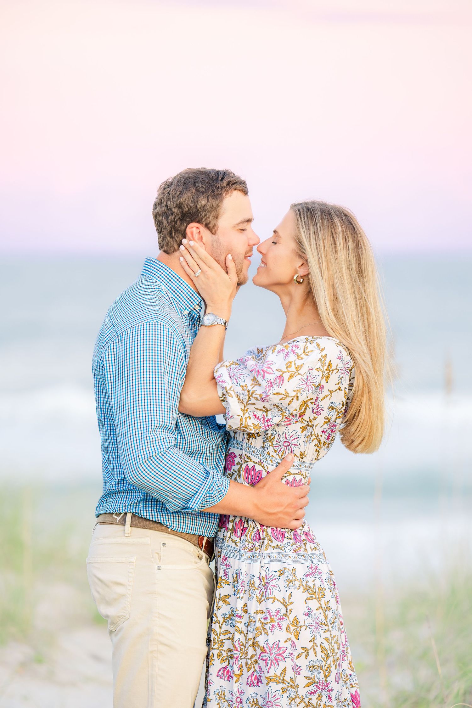 Tender beach moment captured with couple in floral dress and blue shirt against ocean backdrop.