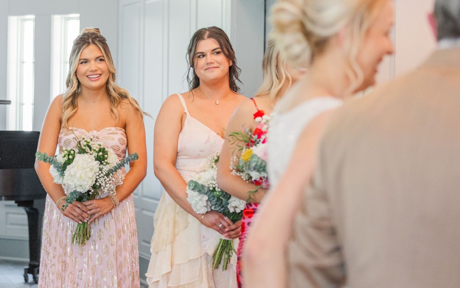 Bridesmaids in blush dresses hold bouquets while standing in elegant white church interior.