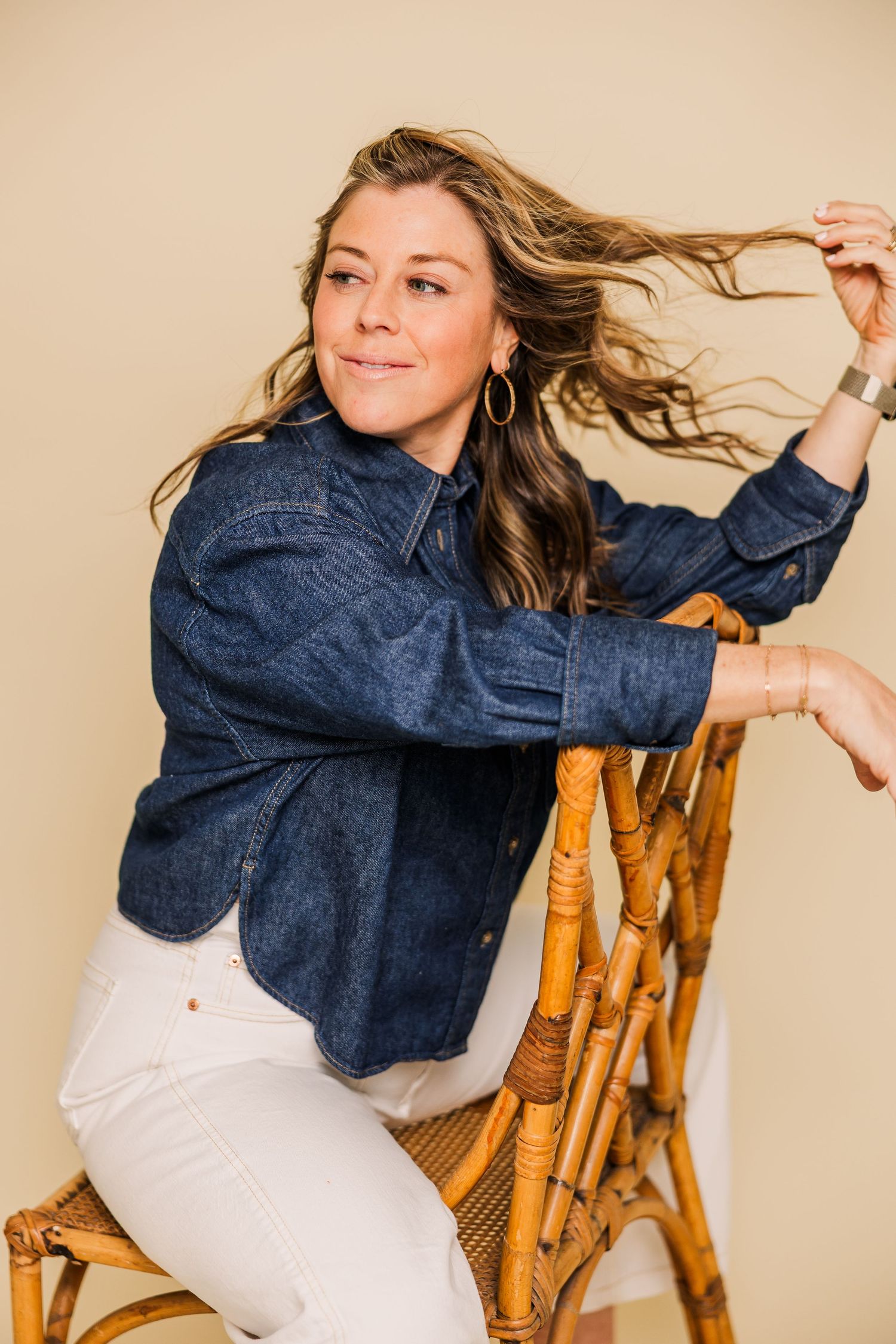 Woman in denim shirt poses playfully while sitting on bamboo chair against beige background.