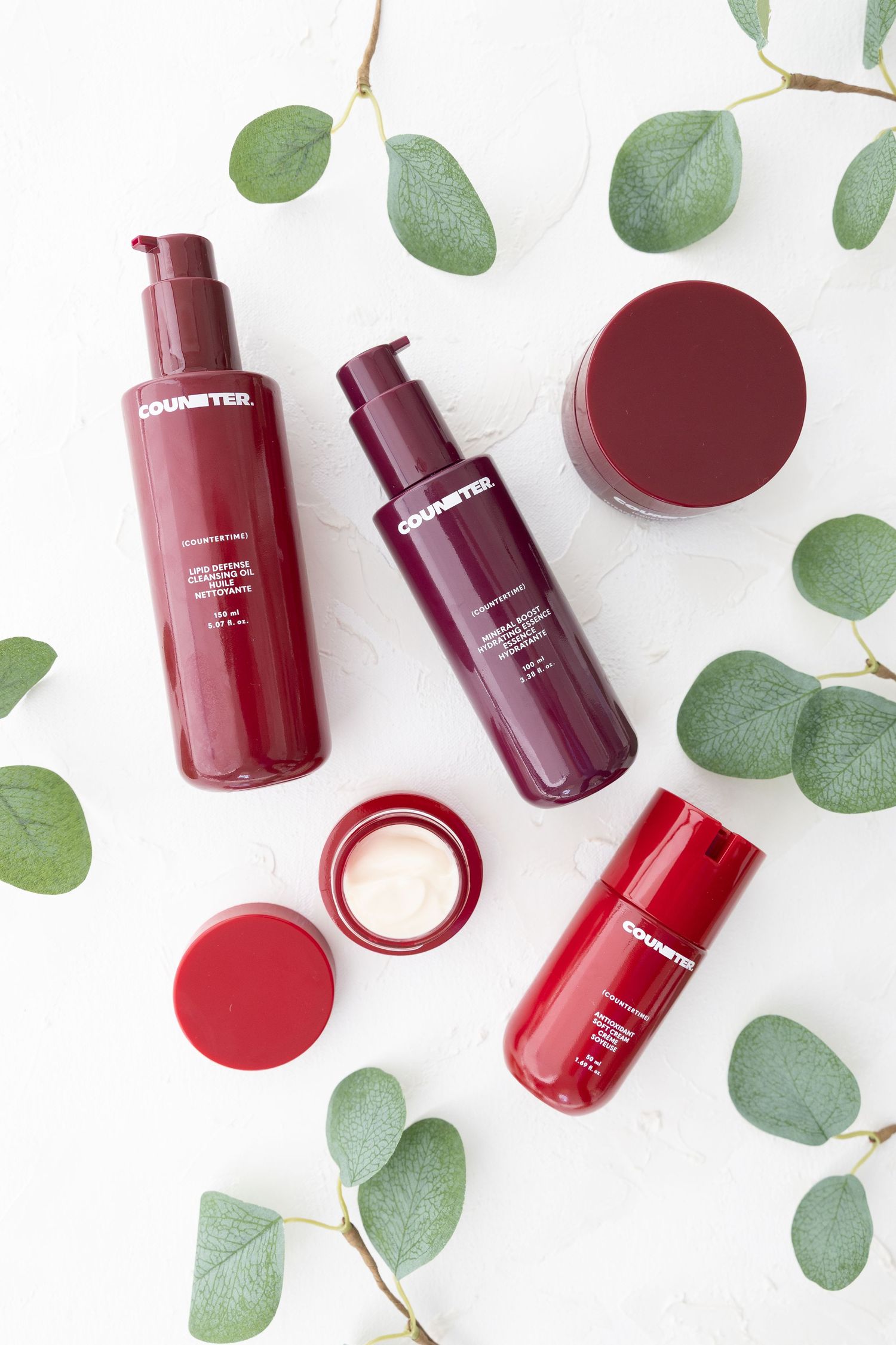 Collection of burgundy skincare bottles arranged with eucalyptus leaves on white background.