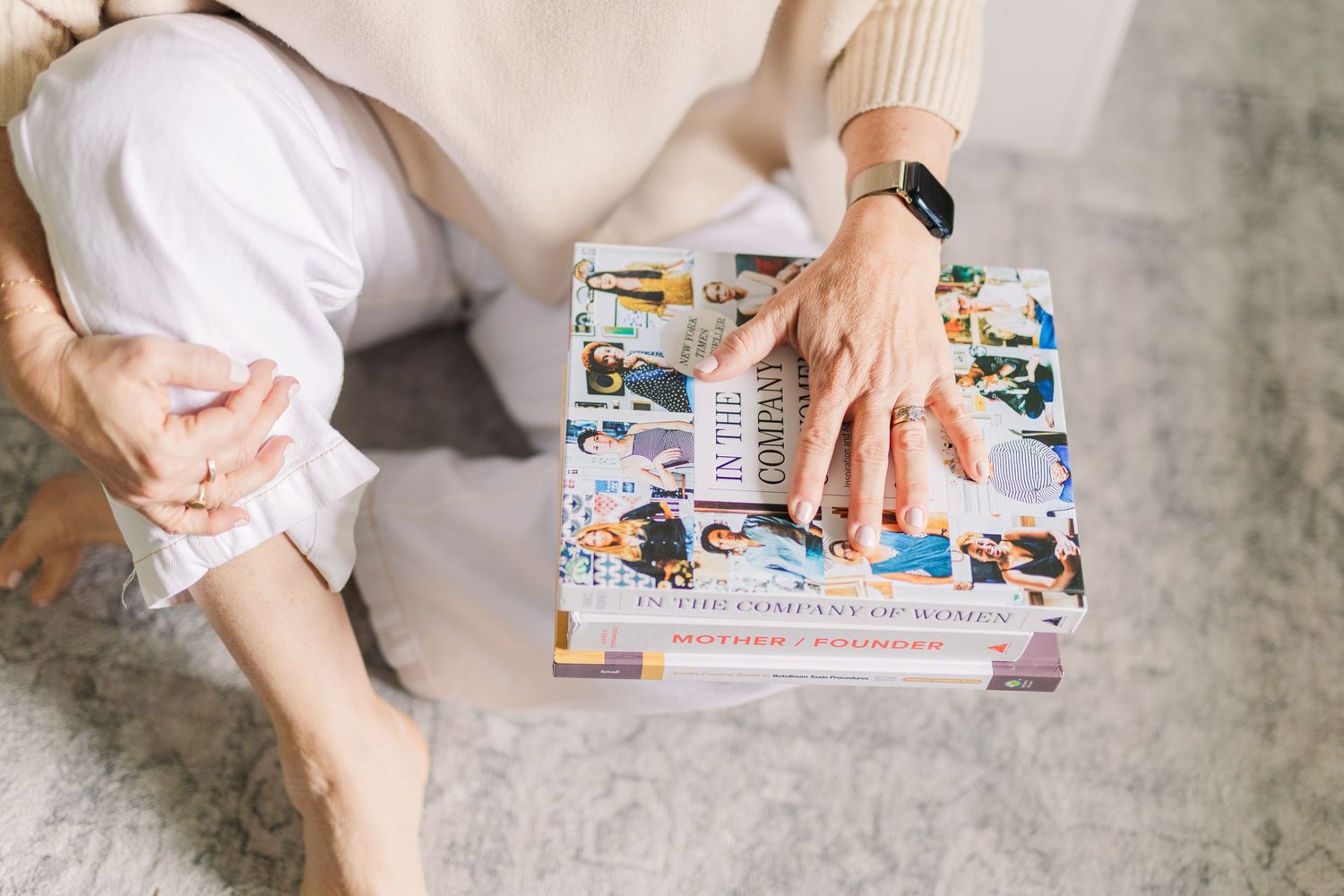 Detail shot of hands resting on stack of magazines while wearing cream sweater and white pants.