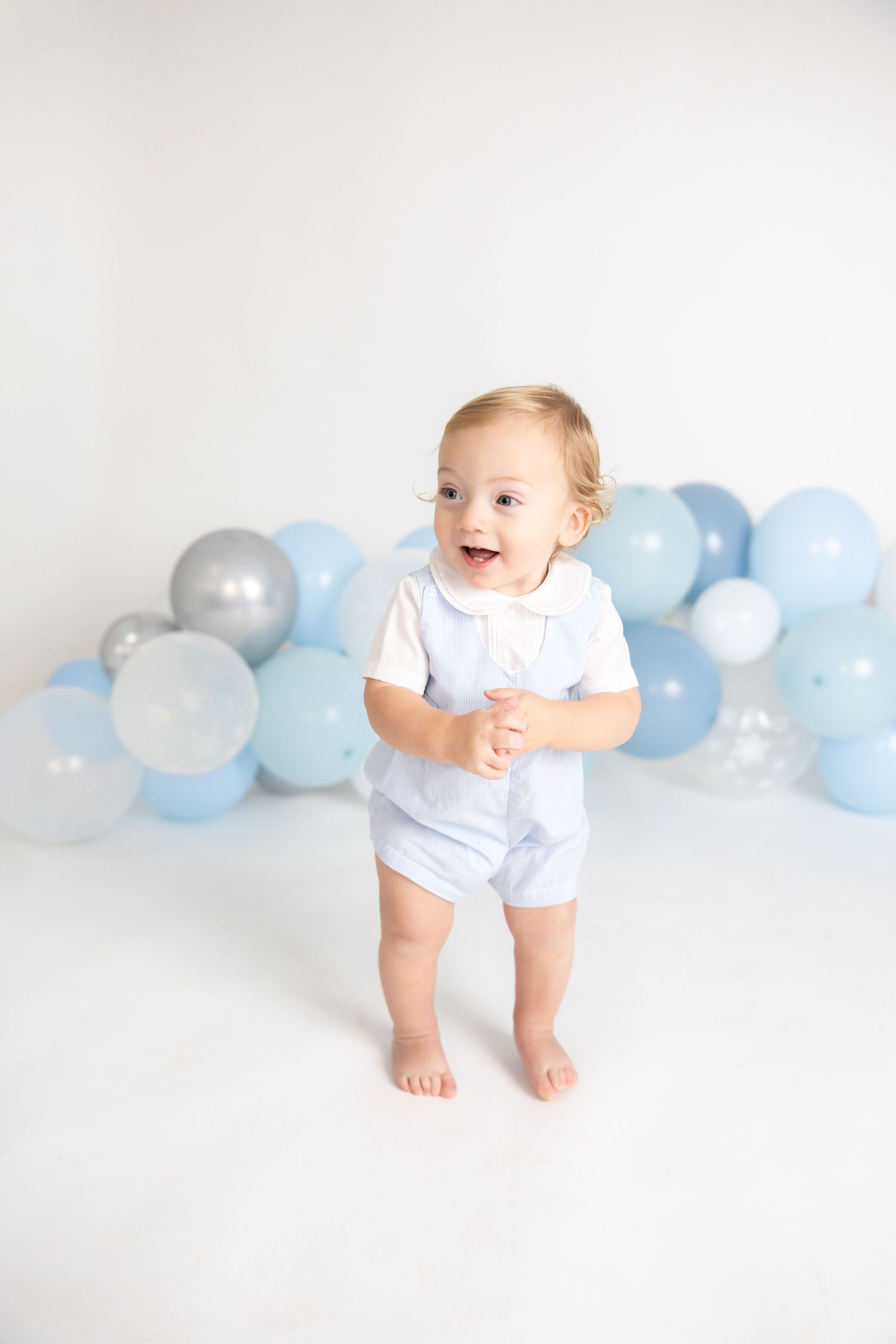 Baby wearing white outfit crawling on floor with pastel blue balloon garland backdrop.