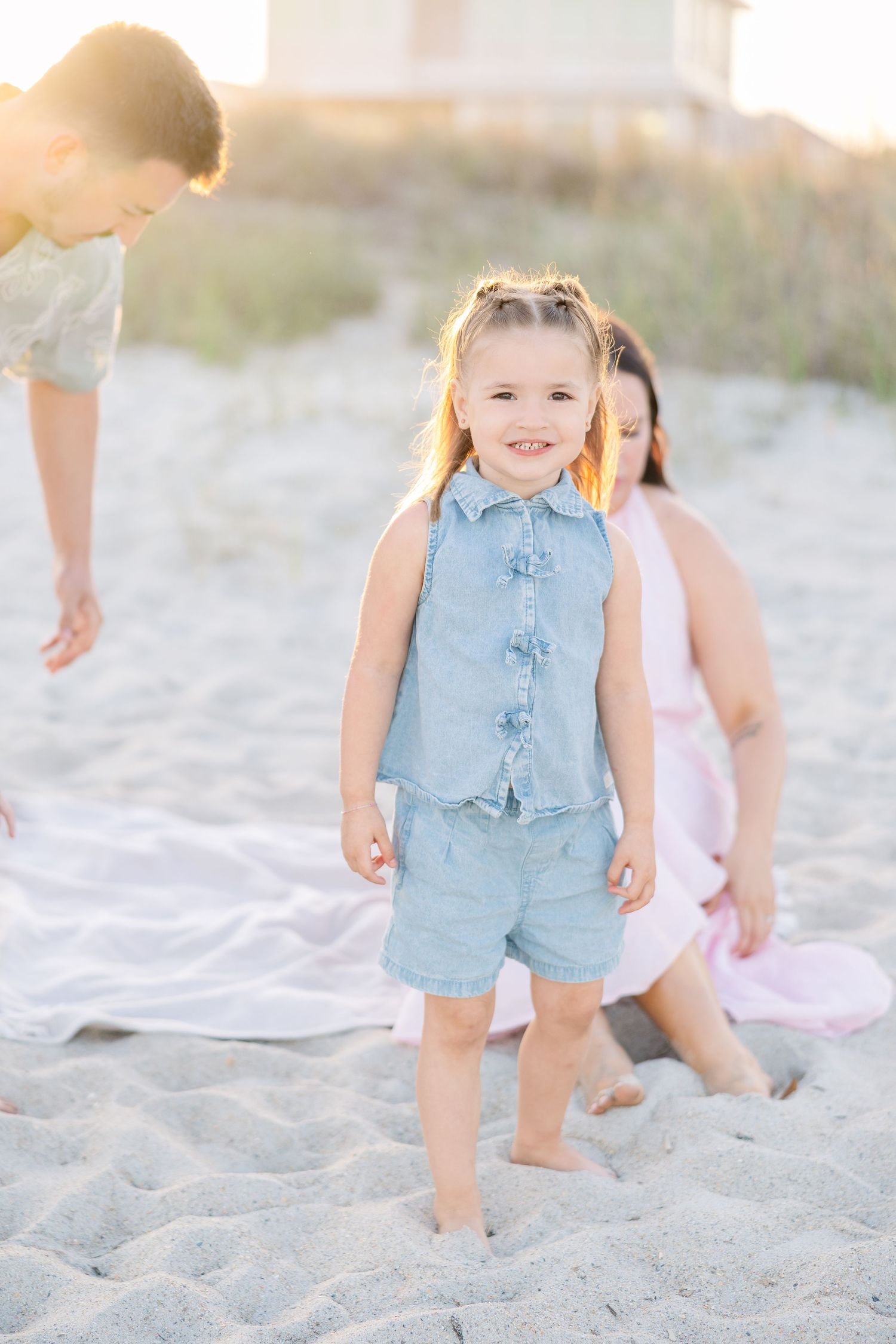 A smiling toddler girl in a denim outfit stands barefoot on sandy beach with parents in soft sunlight.