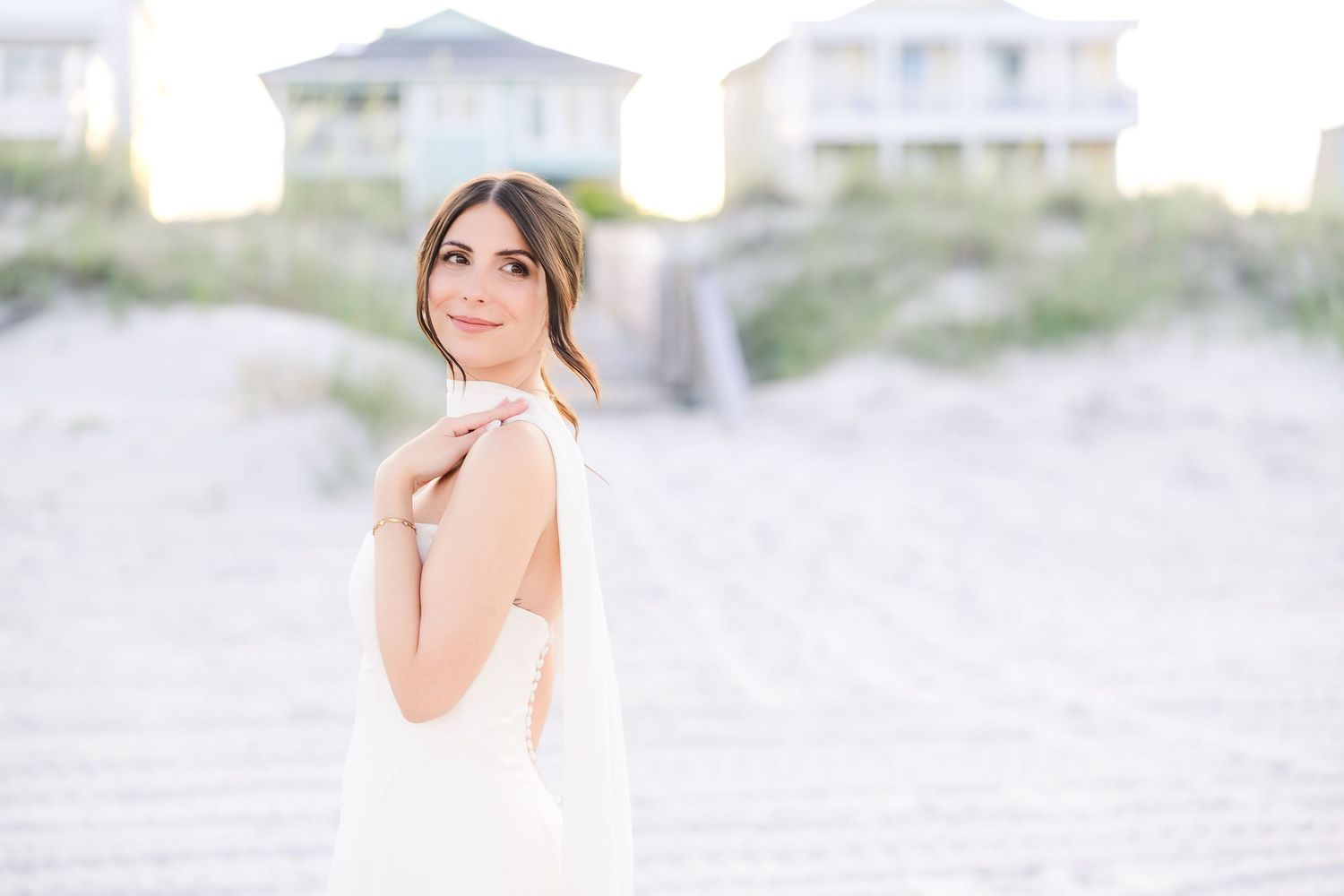 Bride in white halter dress poses on sandy beach with coastal homes in background in Carolina Beach near Wilmington, NC.