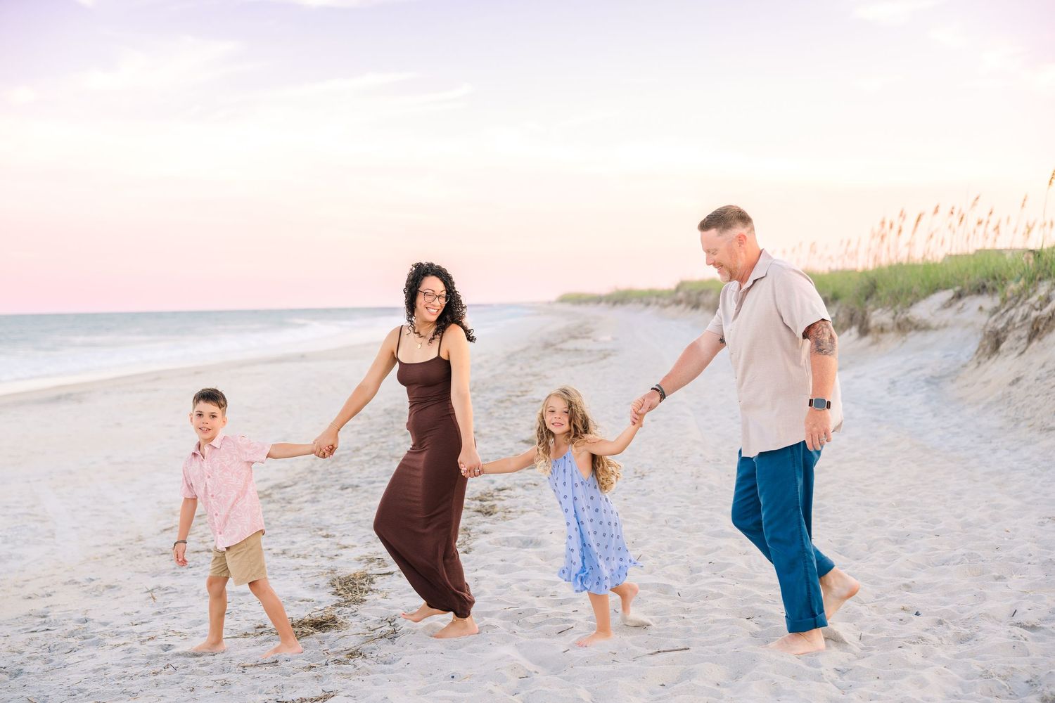 Family of four holding hands and walking on a sandy beach at sunset with ocean waves in the background.