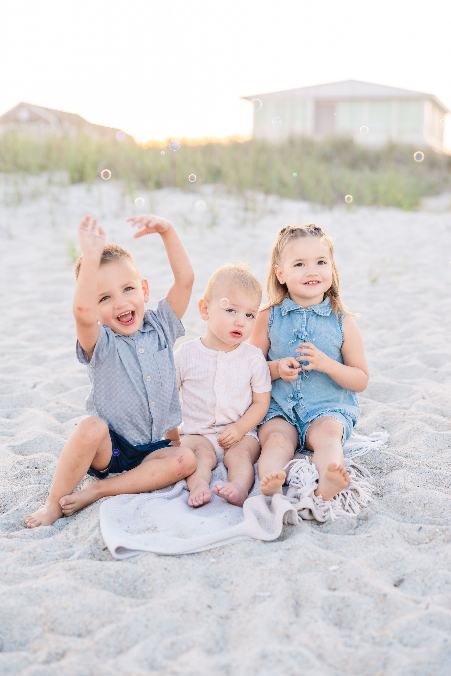 Three young children sit on a white blanket on a sandy beach, playing with bubbles in soft golden light.