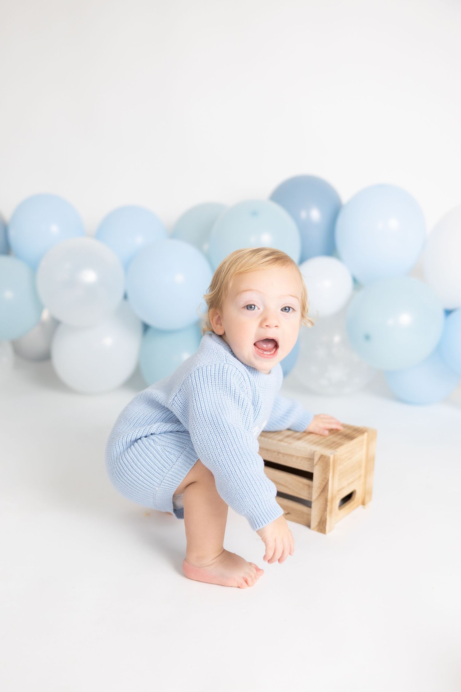 Baby in blue outfit plays near wooden crate with balloon backdrop in studio setting.