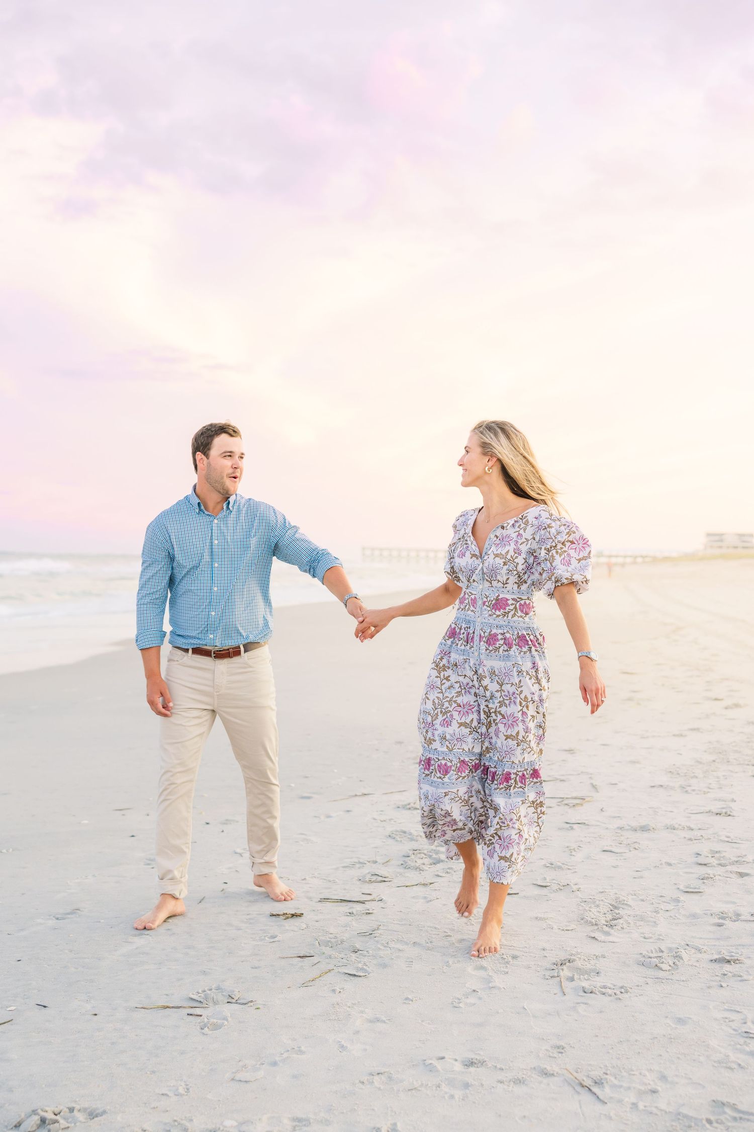 A couple walks hand in hand on a serene beach during sunset with soft pink skies.