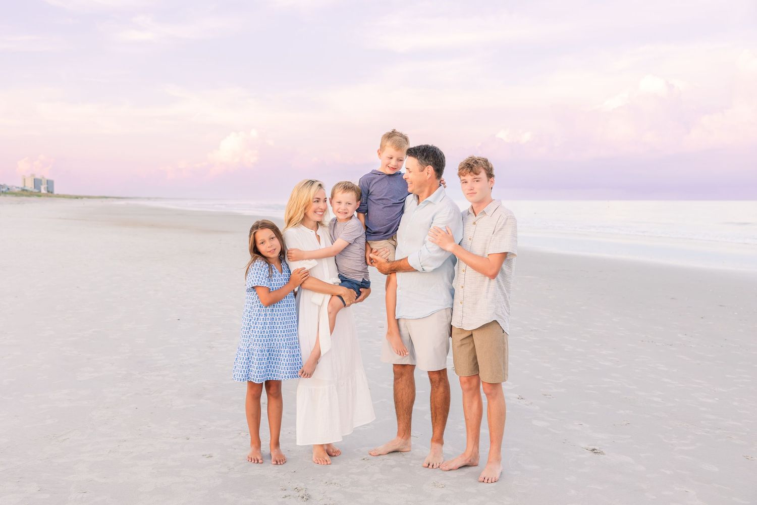 Family group photo on a beach at sunset with soft pink sky in the background.