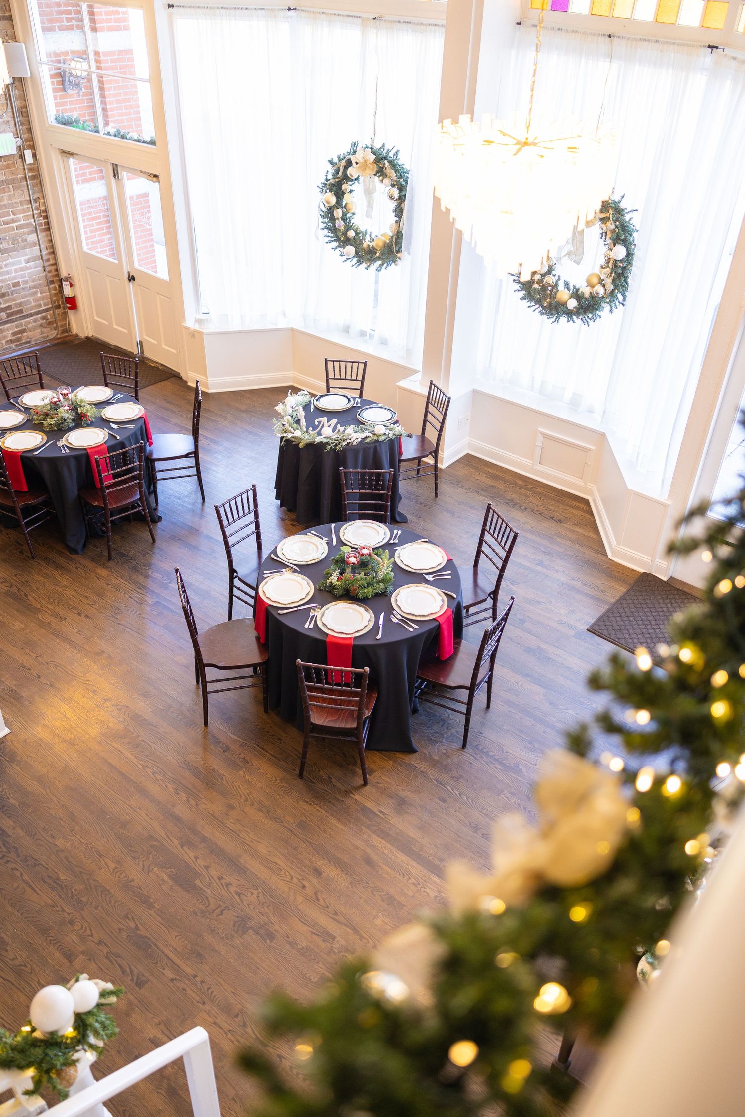 An elegant reception hall with round tables, dark chairs, and holiday wreaths on the windows.