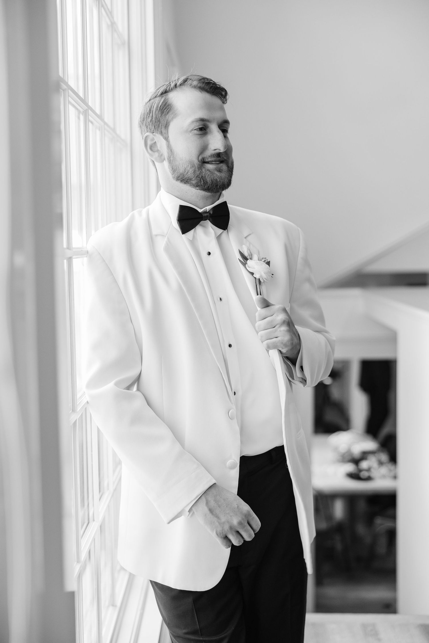 Groom in white tuxedo jacket and black bow tie stands by window, holding boutonniere in black and white photo.