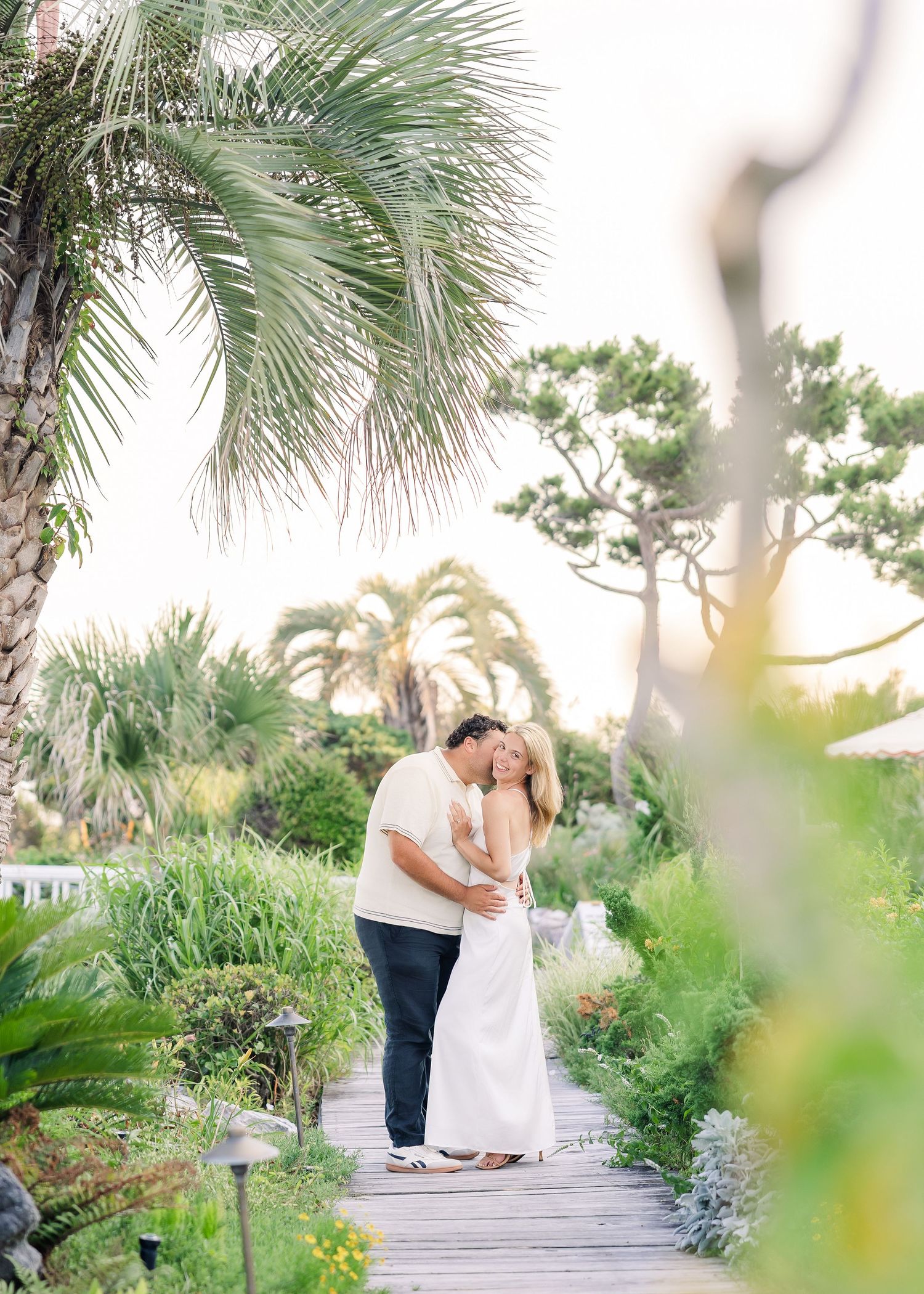 Couple sharing a romantic kiss on a wooden boardwalk surrounded by lush tropical gardens and palm trees.