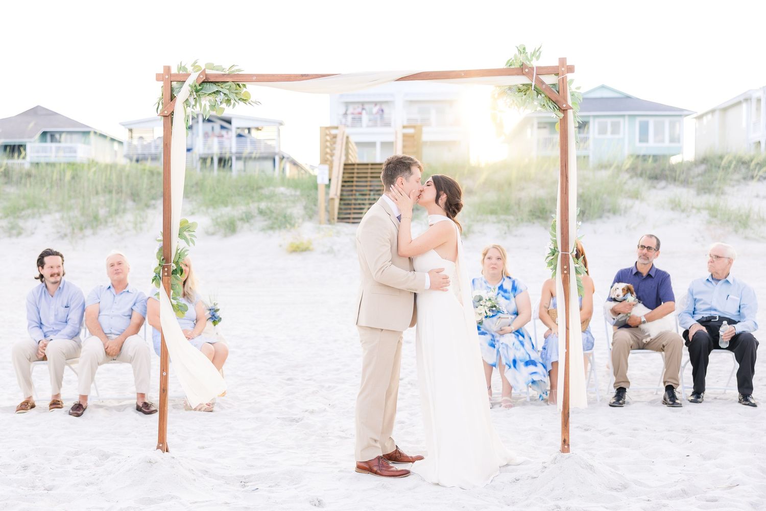Couple shares first kiss at beach wedding ceremony under wooden arch with seated guests on sandy shore in Carolina Beach near Wilmington, NC