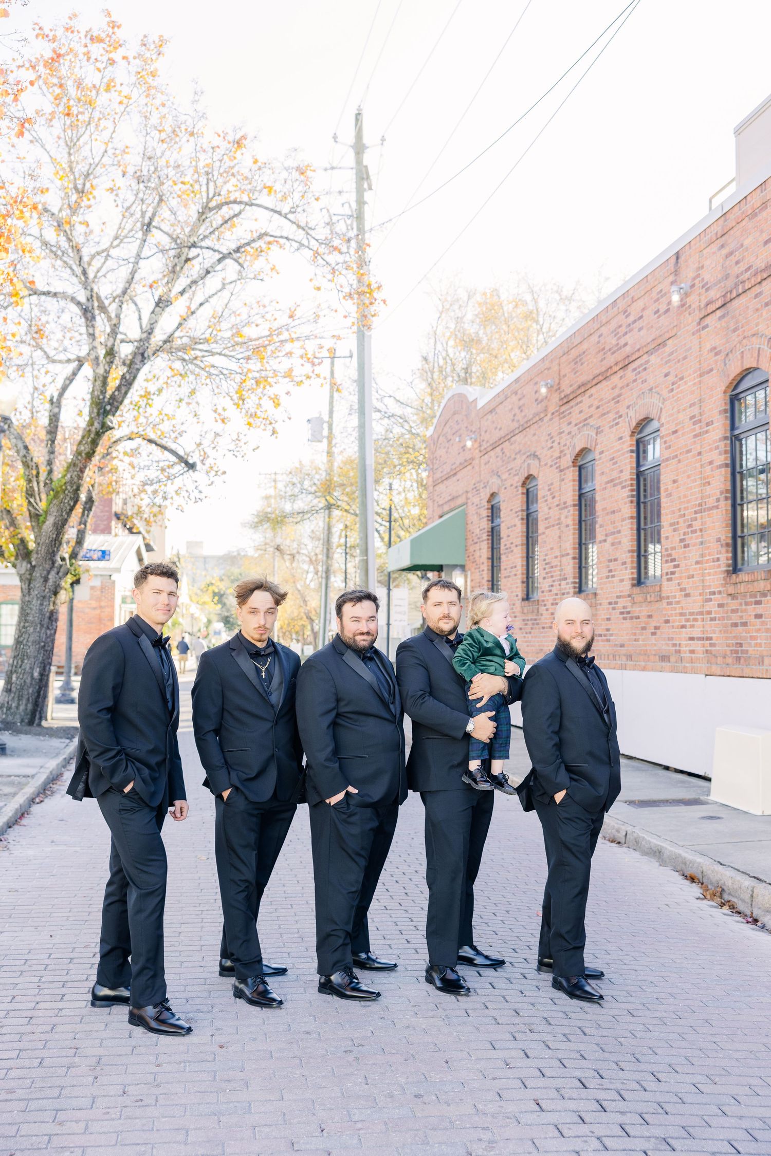 Groomsmen in black suits pose casually on brick sidewalk during wedding photos wedding groom bride wilmington North Carolina photographer