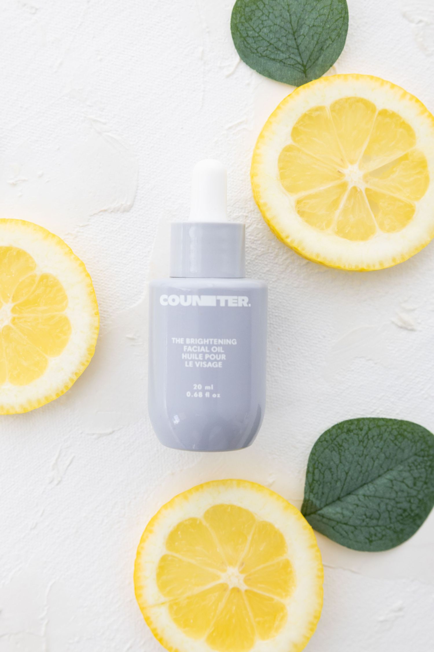 Grey cosmetic bottle with dropper surrounded by fresh lemon slices and green leaves.
