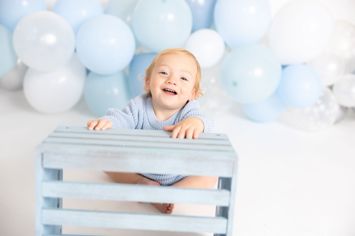 Happy baby plays peek-a-boo with blue wooden crate and balloon background.