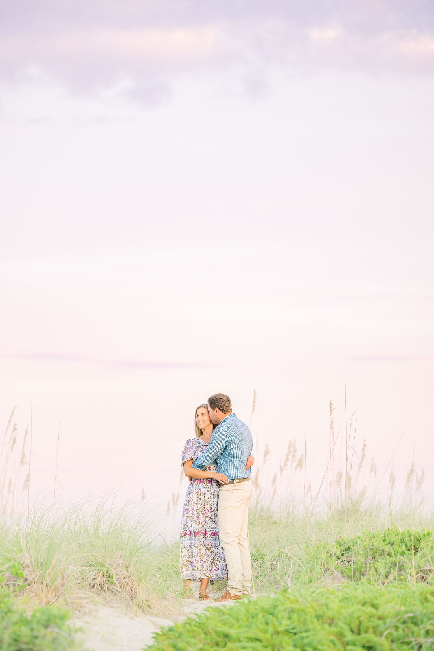 Couple embracing in a field of tall grass during golden hour with ocean in background.