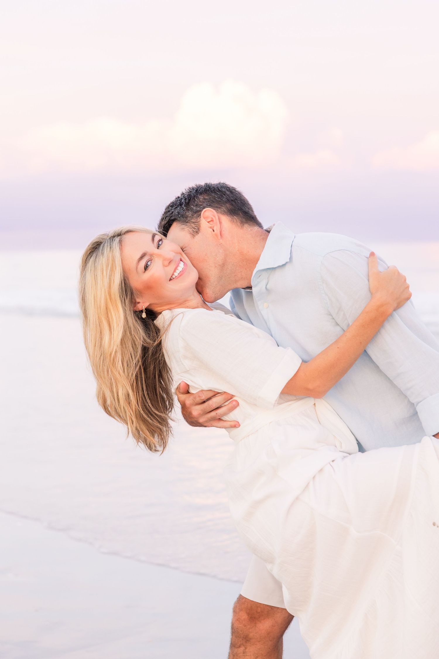 A couple in white clothing shares an intimate dance pose on the beach at dusk.