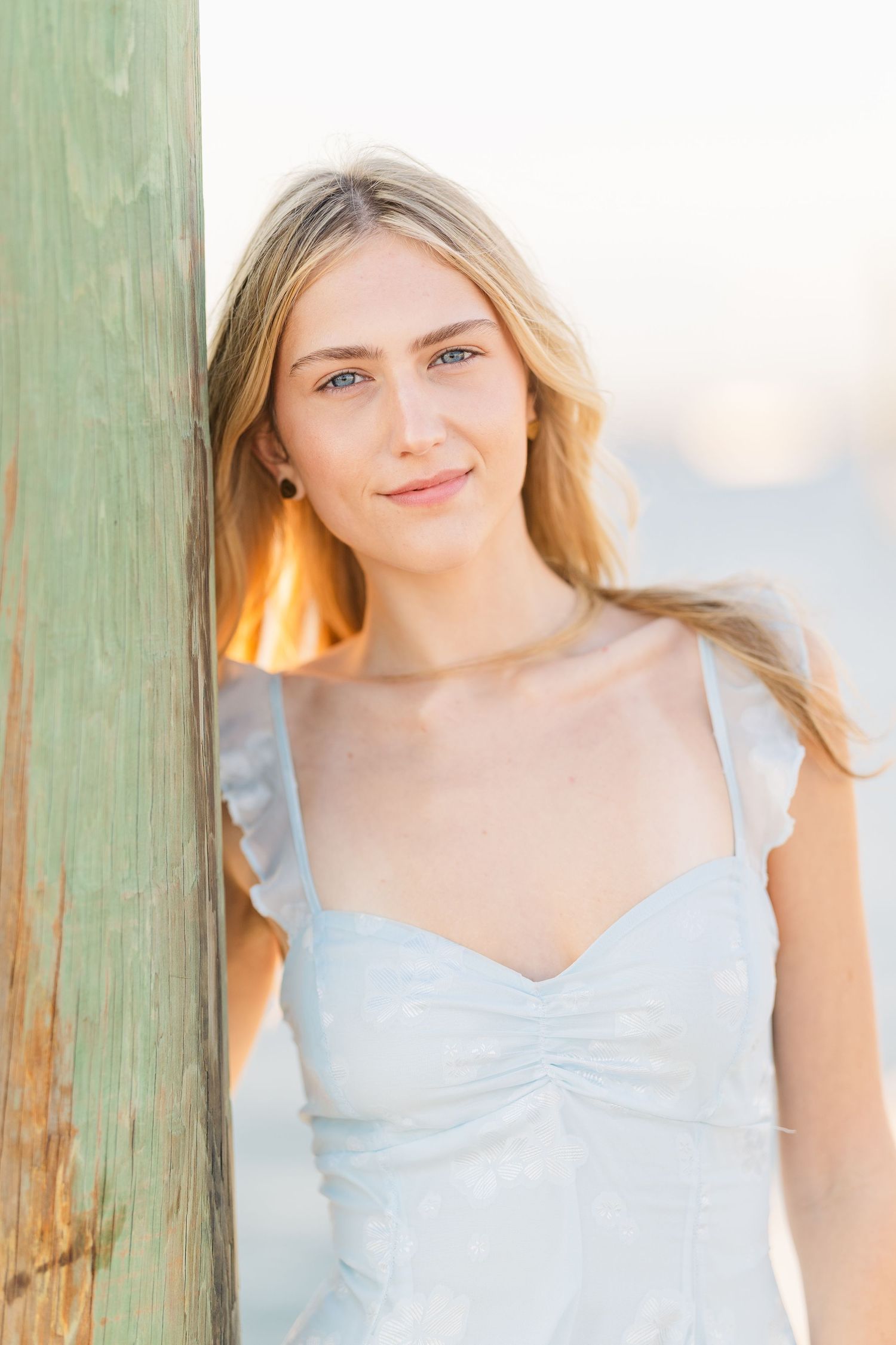 Dreamy portrait in soft light against green post wearing light blue sundress with delicate straps.