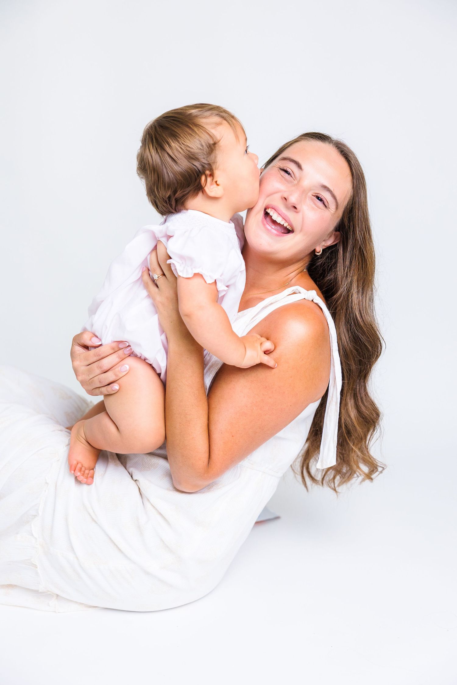 Happy young mother laughing while holding her baby in a white dress against a light background.