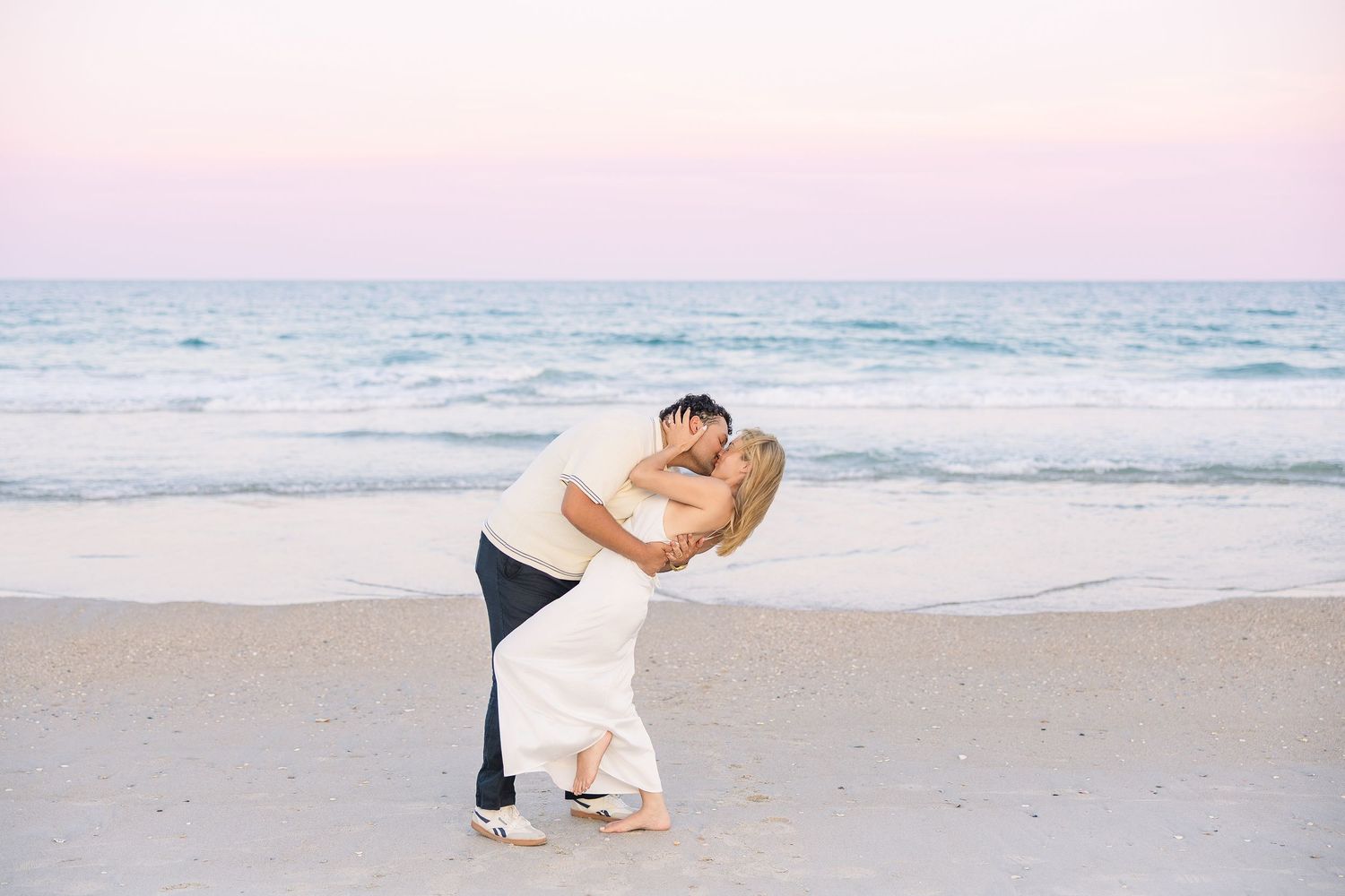 Romantic couple sharing a dip kiss on the beach at sunset with pastel sky backdrop.