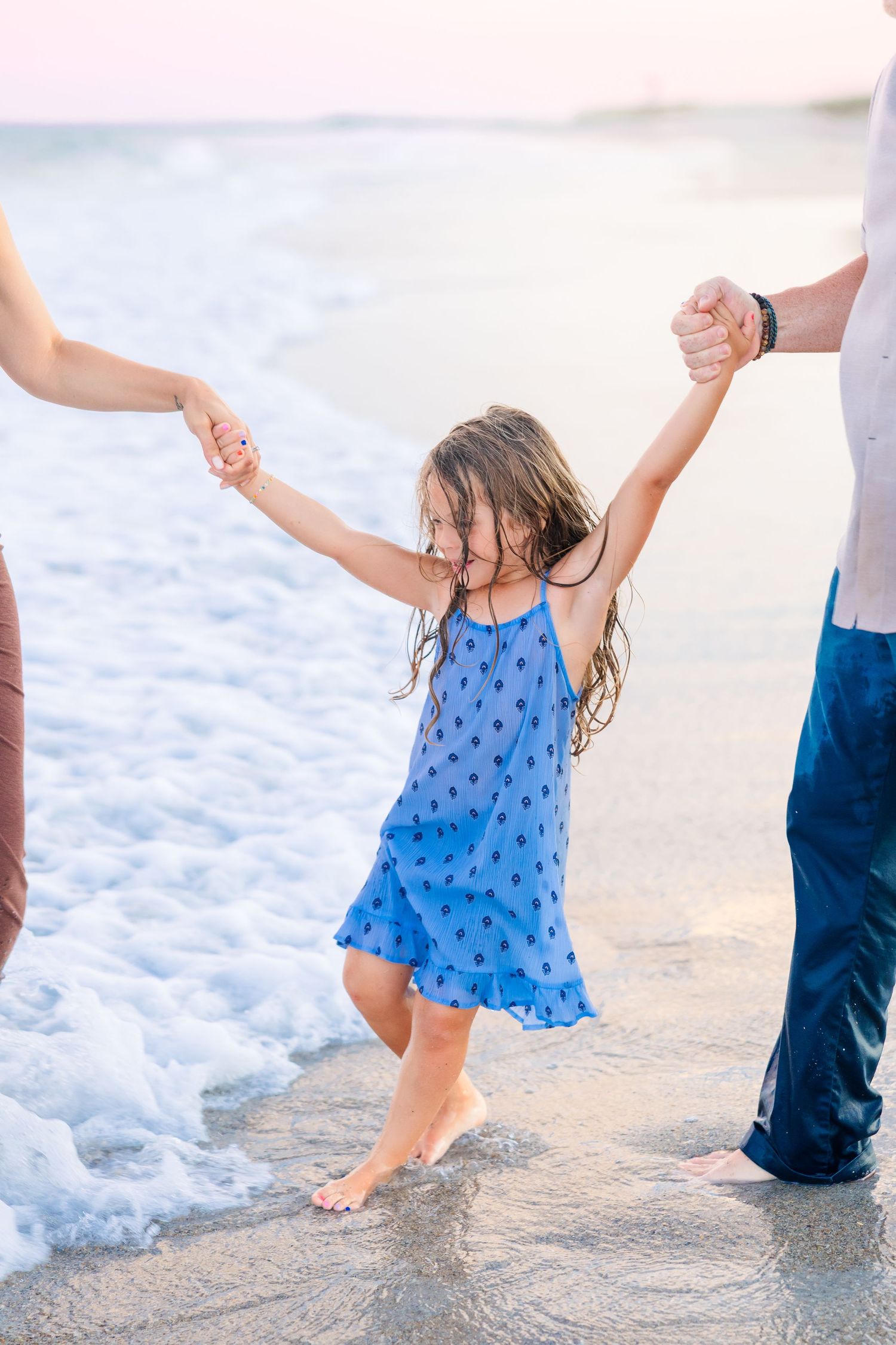 Young girl in blue dress holds parents hands at beach shoreline with ocean waves at sunset.