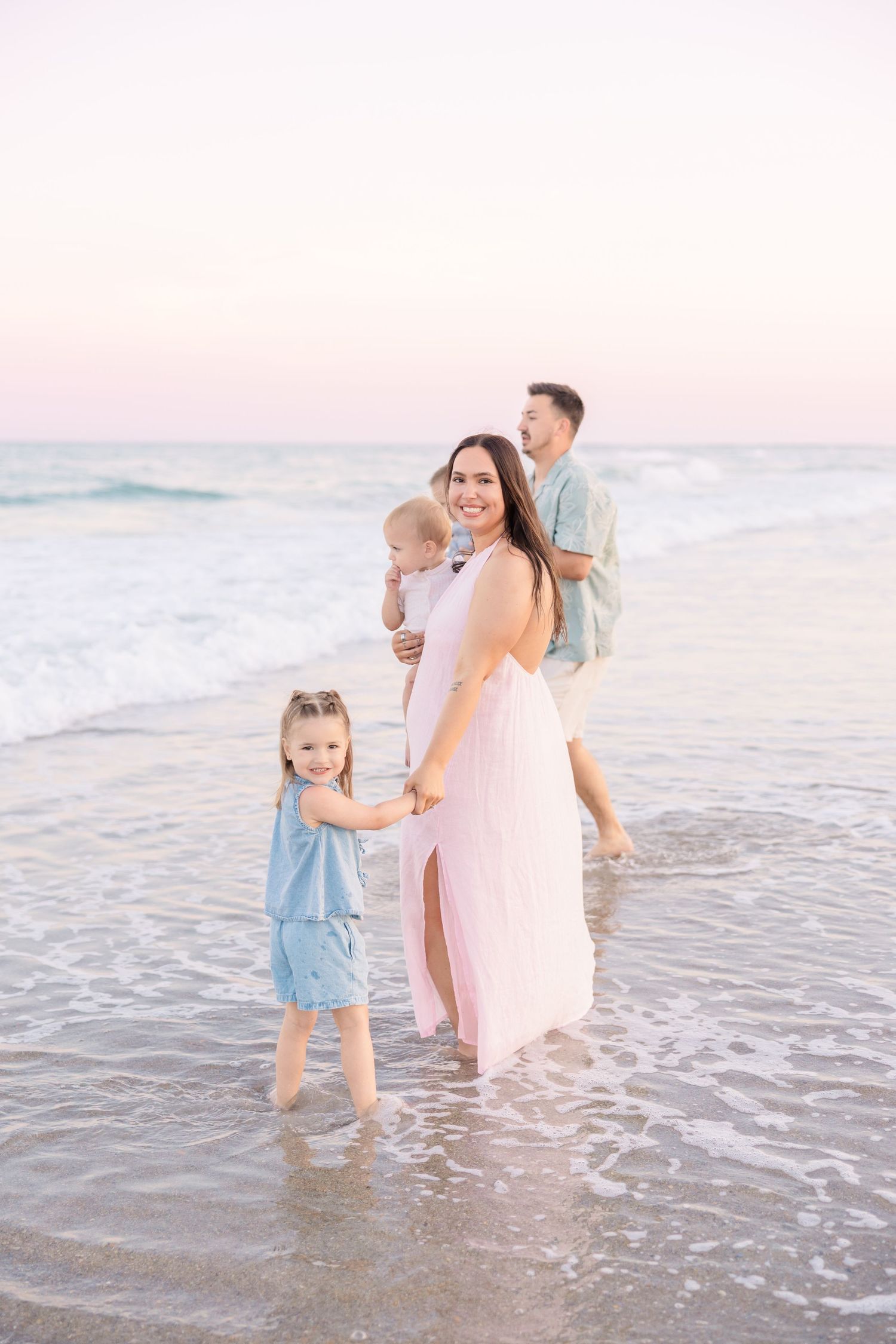 A family of four enjoys a serene beach sunset, mom in pink dress holds toddler while daughter holds her hand.