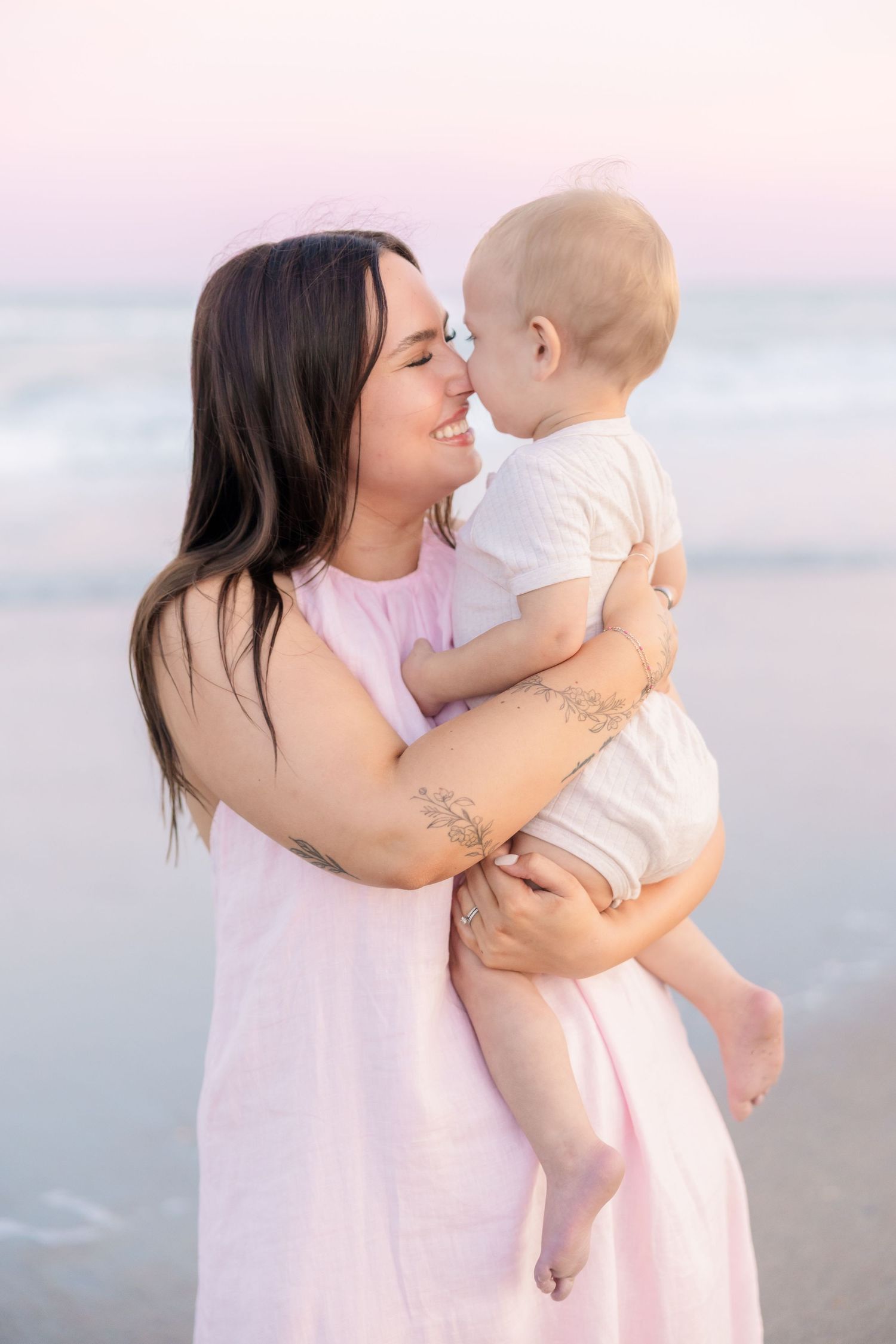 Smiling dark-haired mother in pink dress holds baby close for a nose kiss on a beach at sunset.