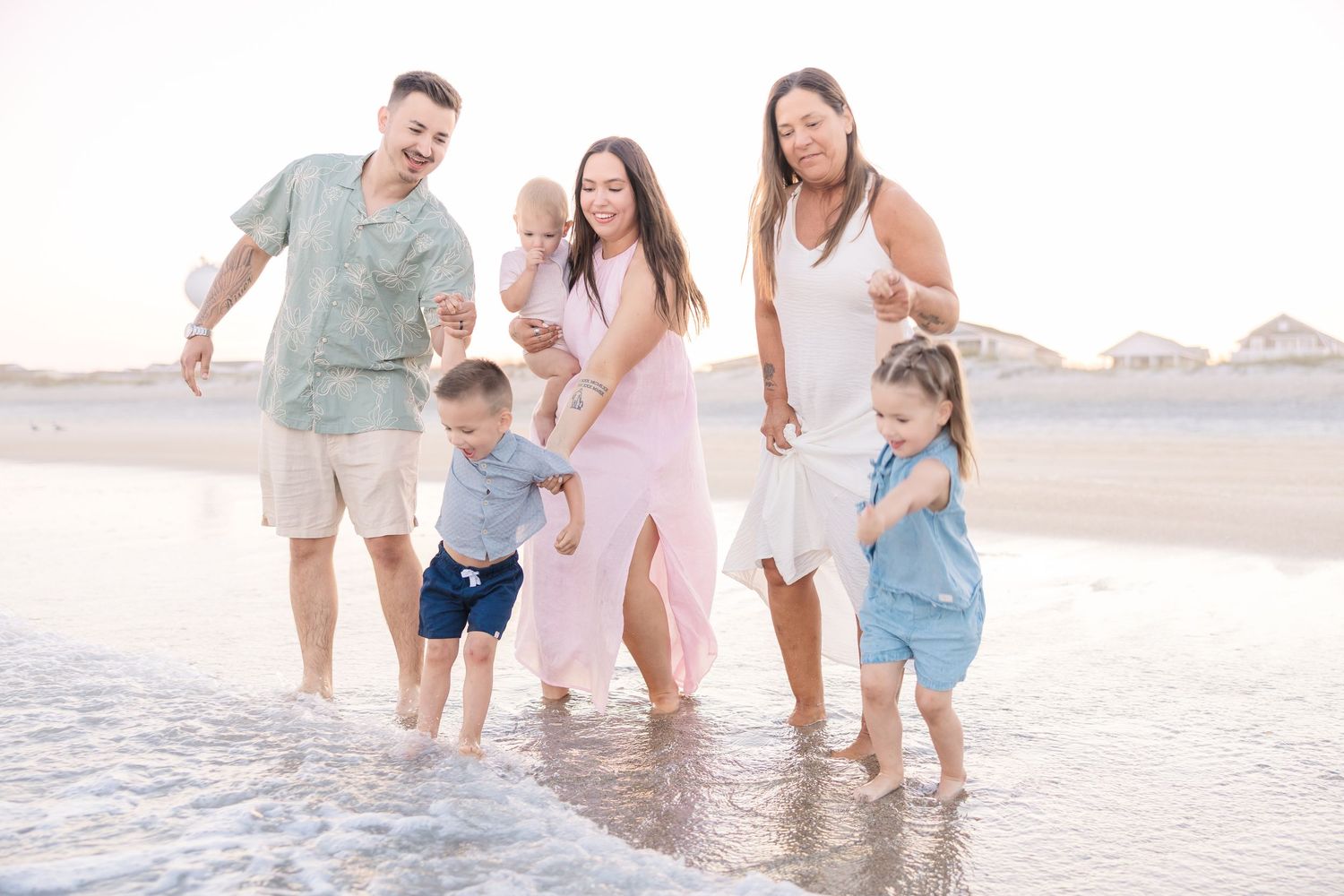 Family of five walking along a sandy beach shore, smiling and enjoying a sunny day together near the ocean waves.