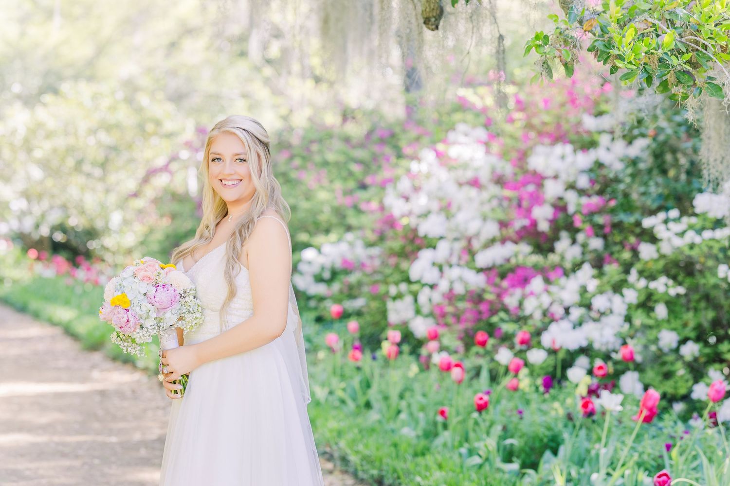 Smiling bride holding colorful bouquet in lush garden with pink and white blooming flowers on sunny day in Airlie Gardens in Wilmington, NC
