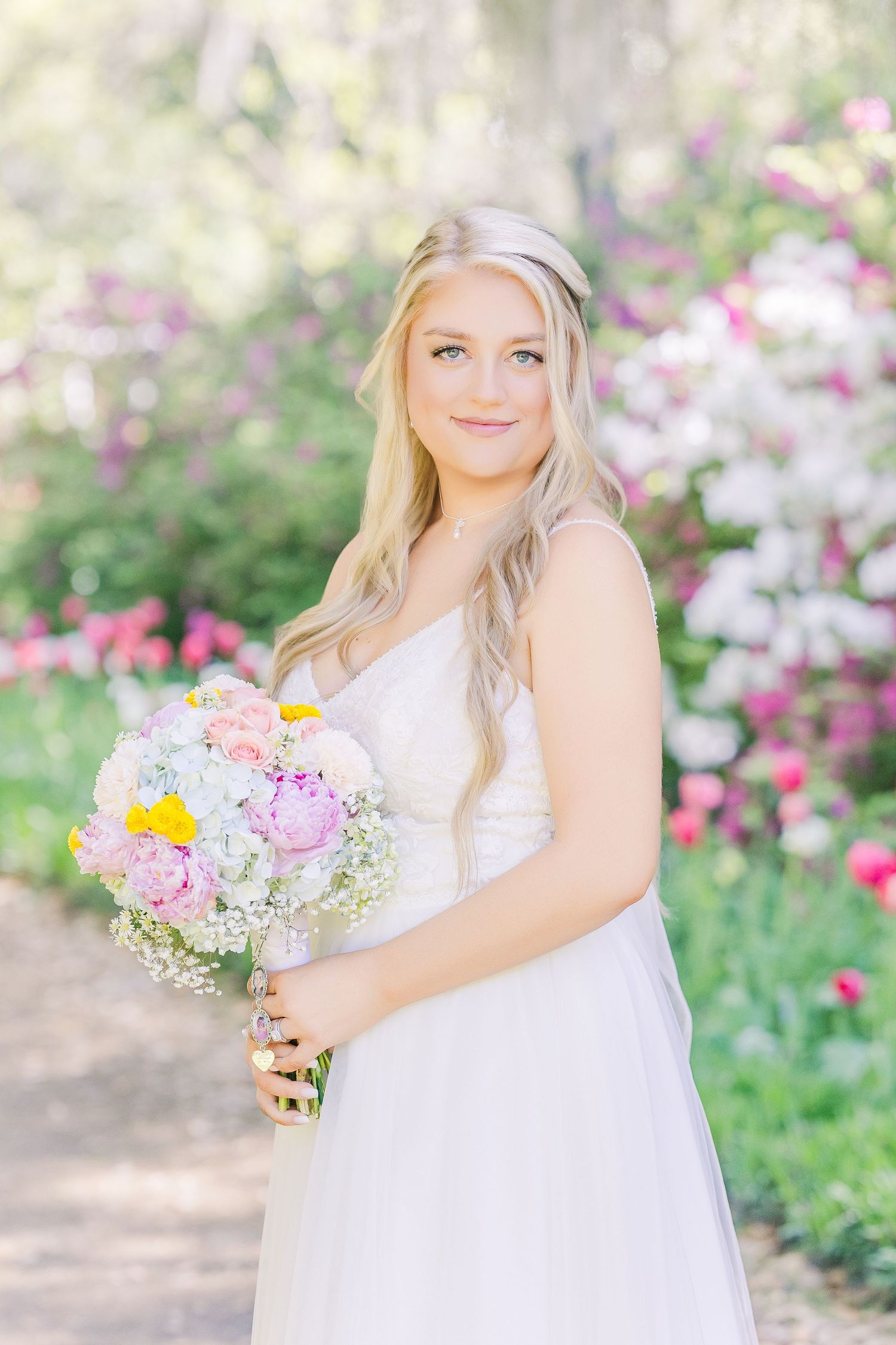 Blonde bride in white gown holds colorful bouquet among blooming garden flowers in Airlie Gardens in Wilmington, NC
