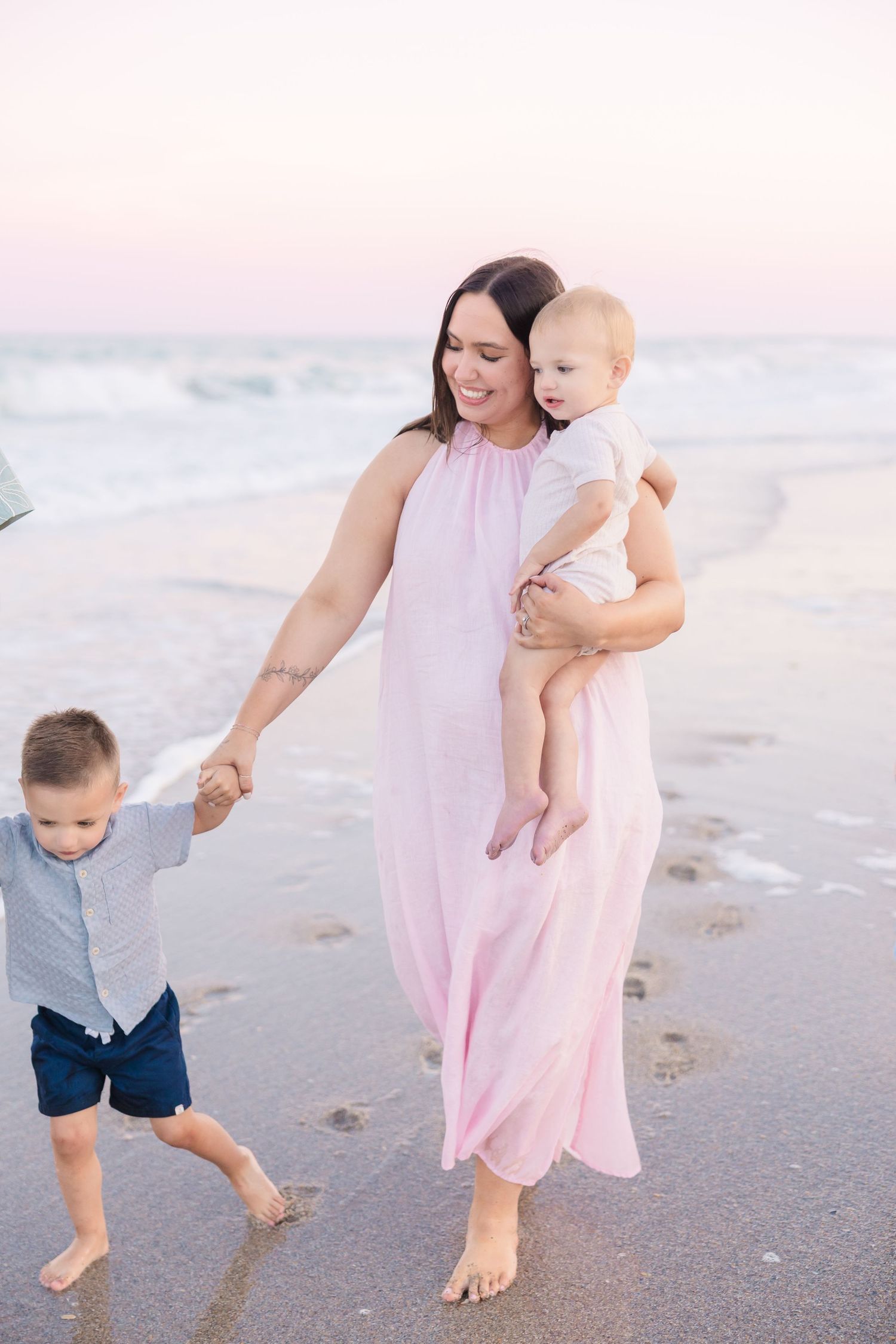 Smiling mother in pink dress carries baby while holding toddler sons hand on sunset beach.