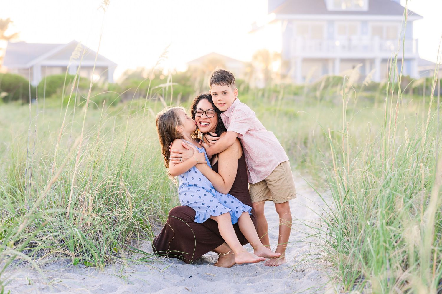 Mom hugging two children in sandy beach dunes at golden hour.