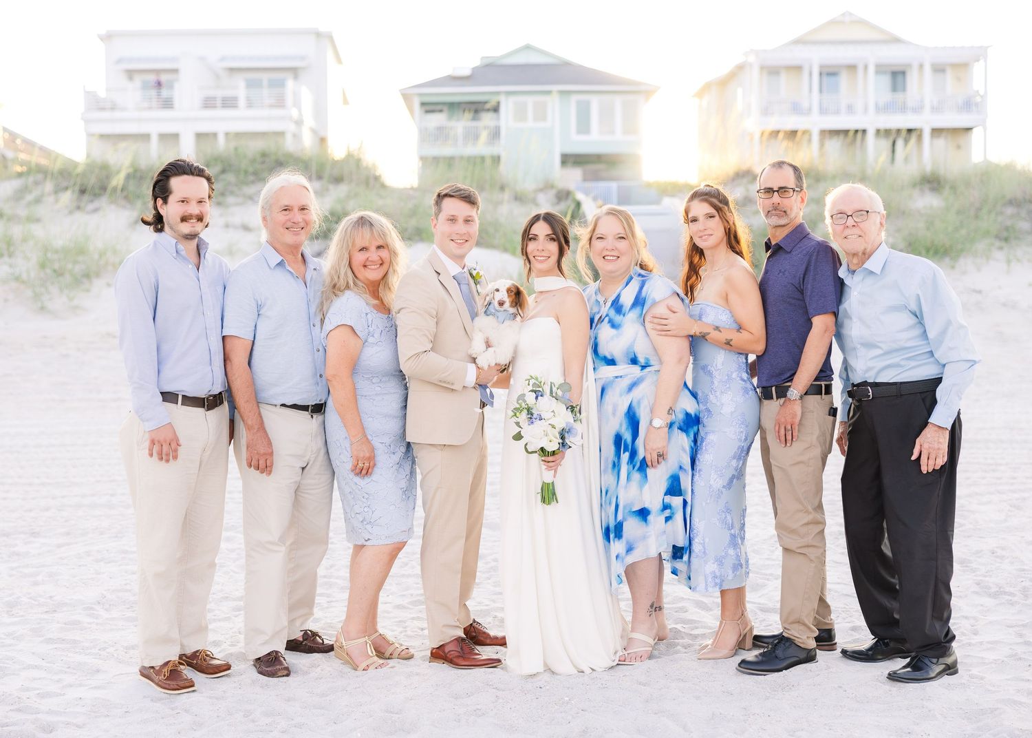Wedding party group photo on sandy beach with coastal homes and dunes in background in Carolina Beach near Wilmington, NC.
