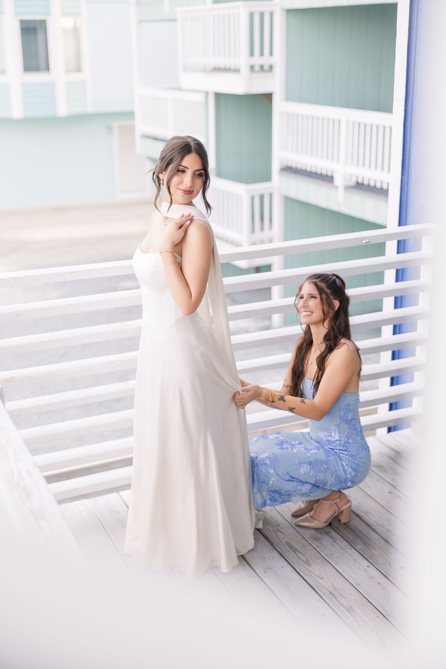 Bridesmaid in blue floral dress adjusts bride's white wedding gown train on a coastal wooden balcony in Carolina Beach near Wilmington, NC.